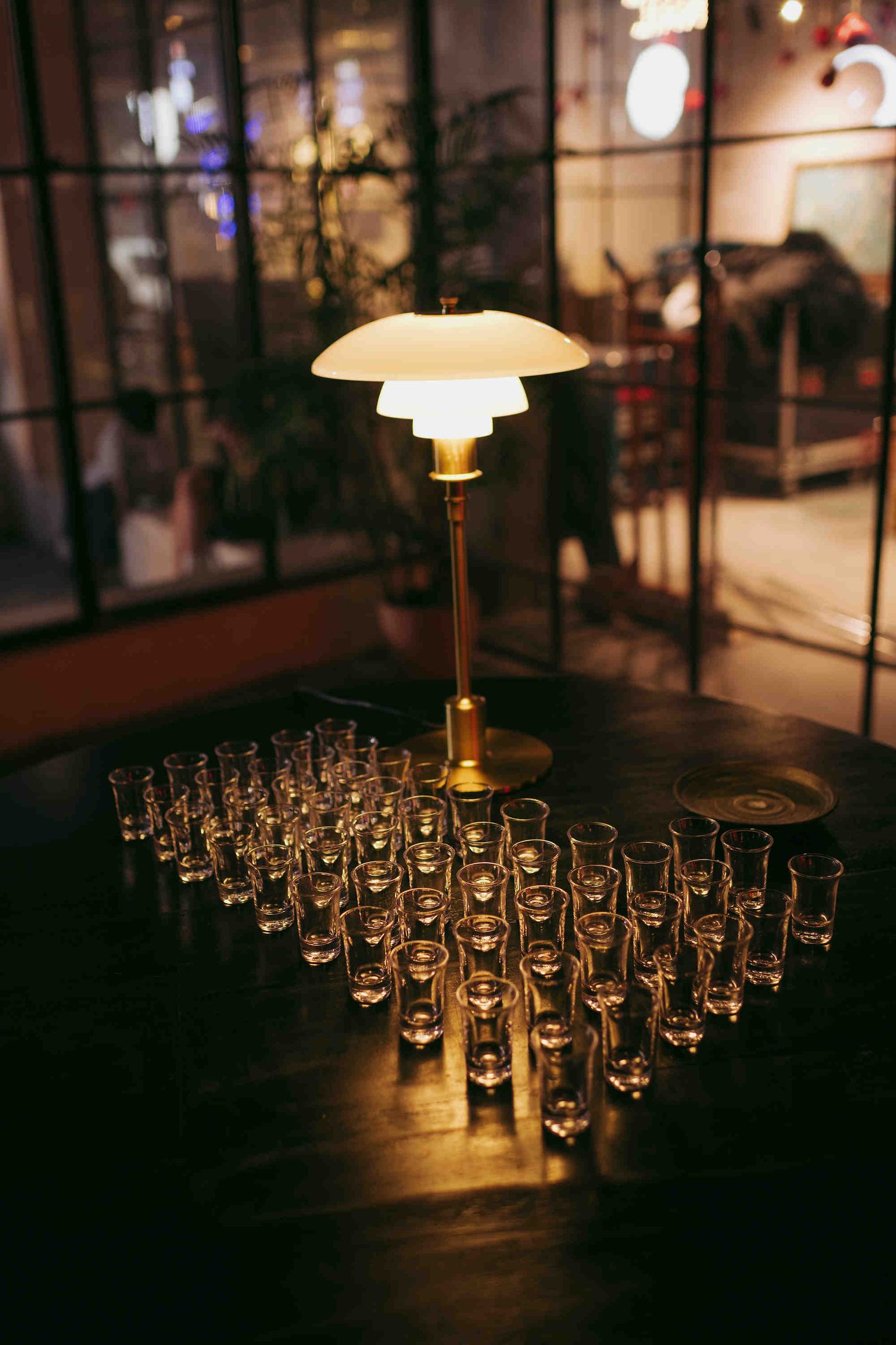 Elegant brass table lamp surrounded by rows of shot glasses on a dark wooden bar table in a dimly lit venue.