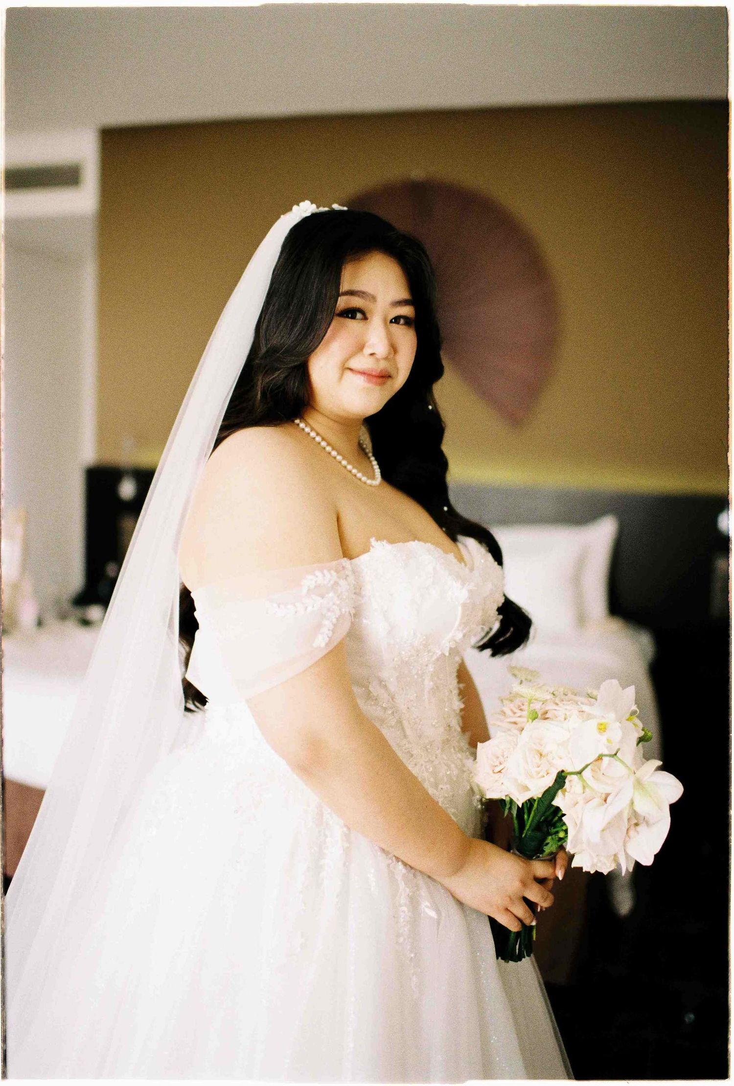 Plus-size bride in off-shoulder white lace wedding gown with veil and pearl necklace holding bouquet of white roses.
