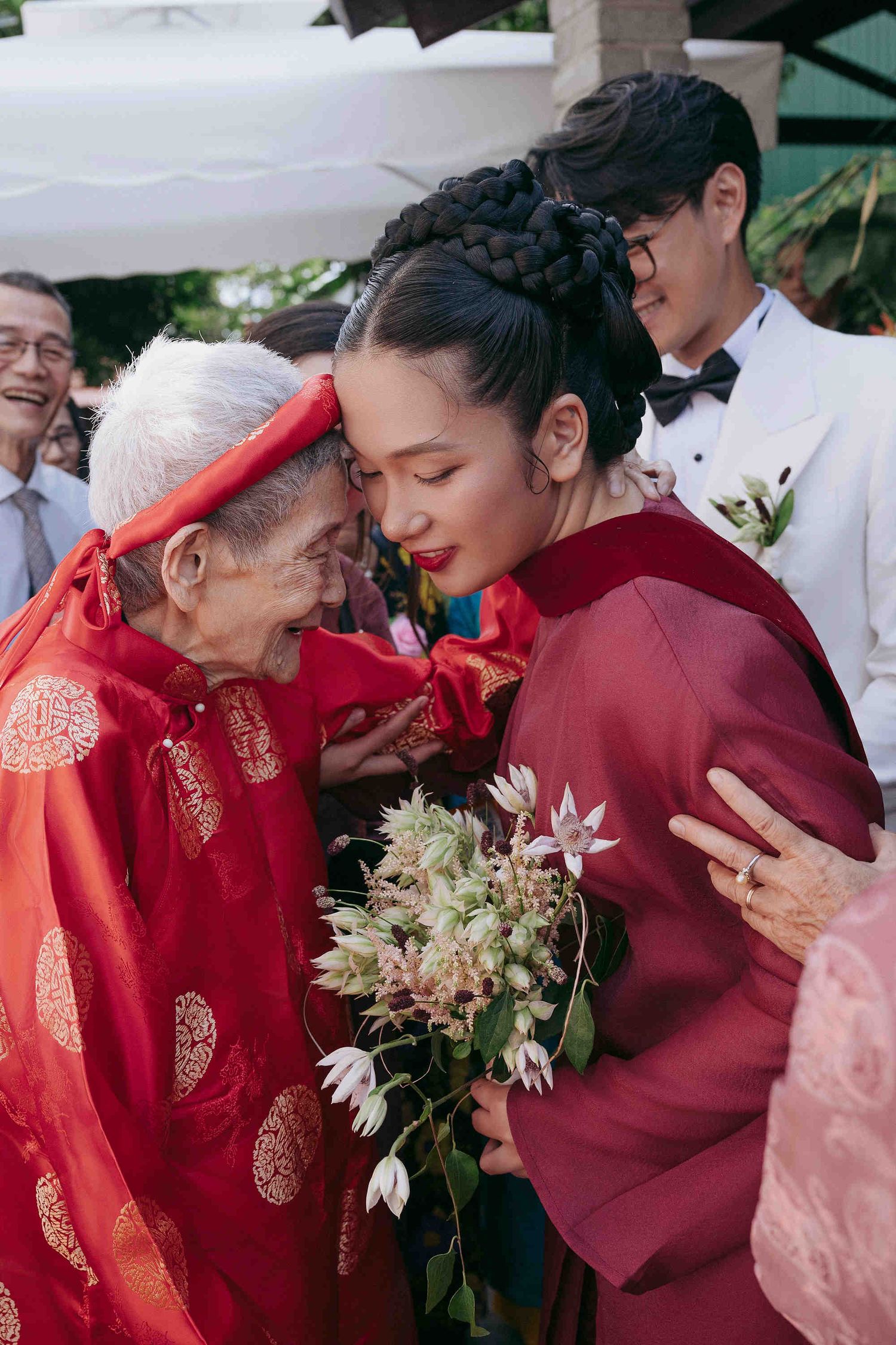 Bride in red ao dai embraces elderly woman in matching red traditional Vietnamese wedding attire while holding a floral bouquet.