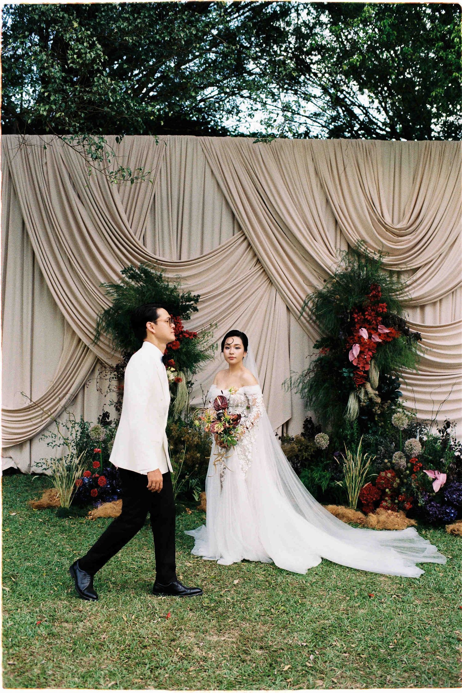 Couple in wedding attire posing before draped cream backdrop with lush floral arrangements at outdoor garden ceremony.