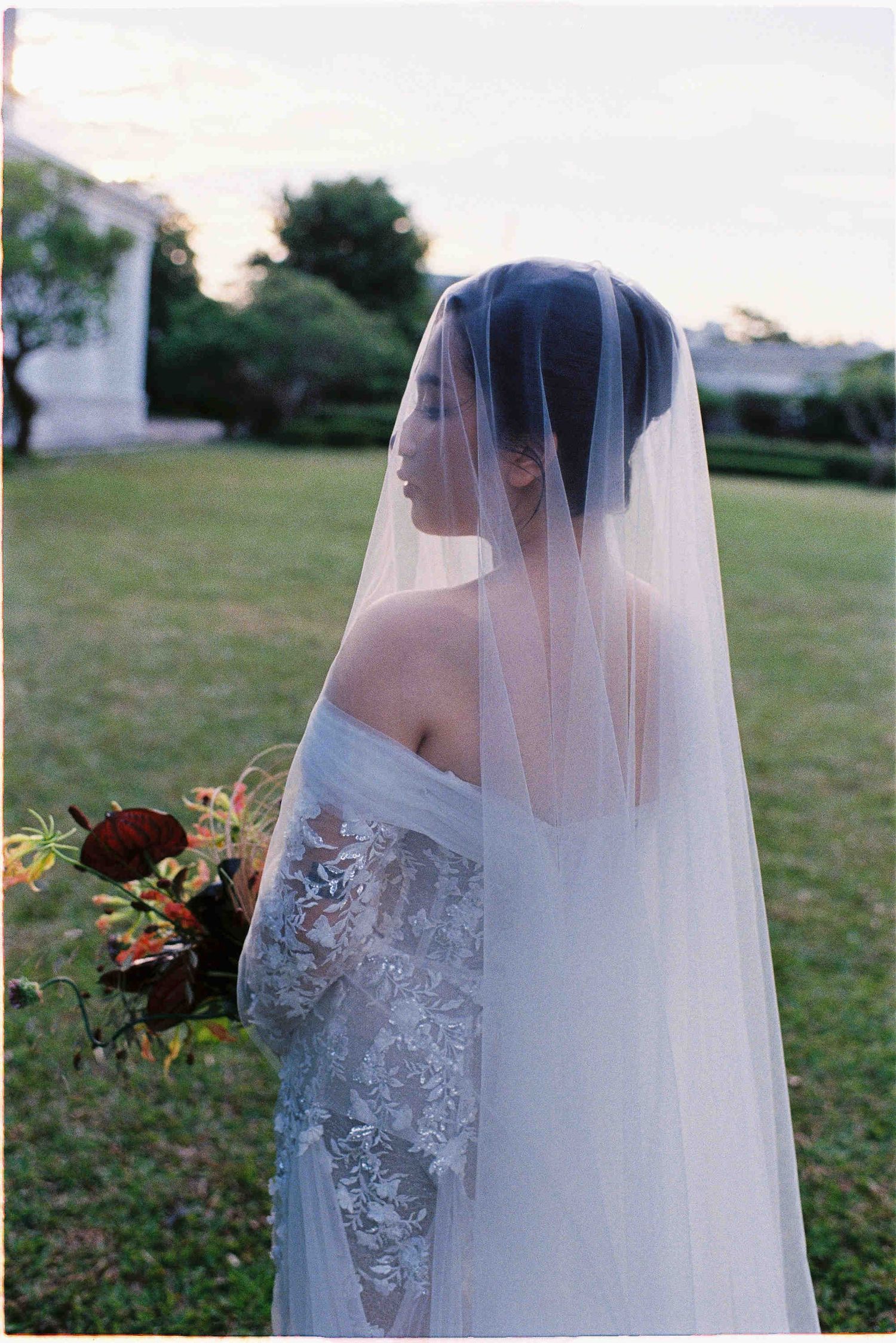 Bride in lace off-shoulder gown and long veil holds dark floral bouquet outdoors at dusk.