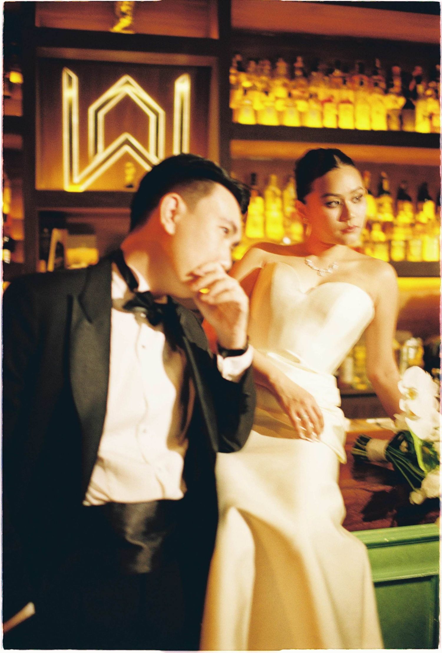 Vietnam pre-wedding: couple in formal attire poses playfully at bar with backlit bottles on display, by AnhVuWedding.