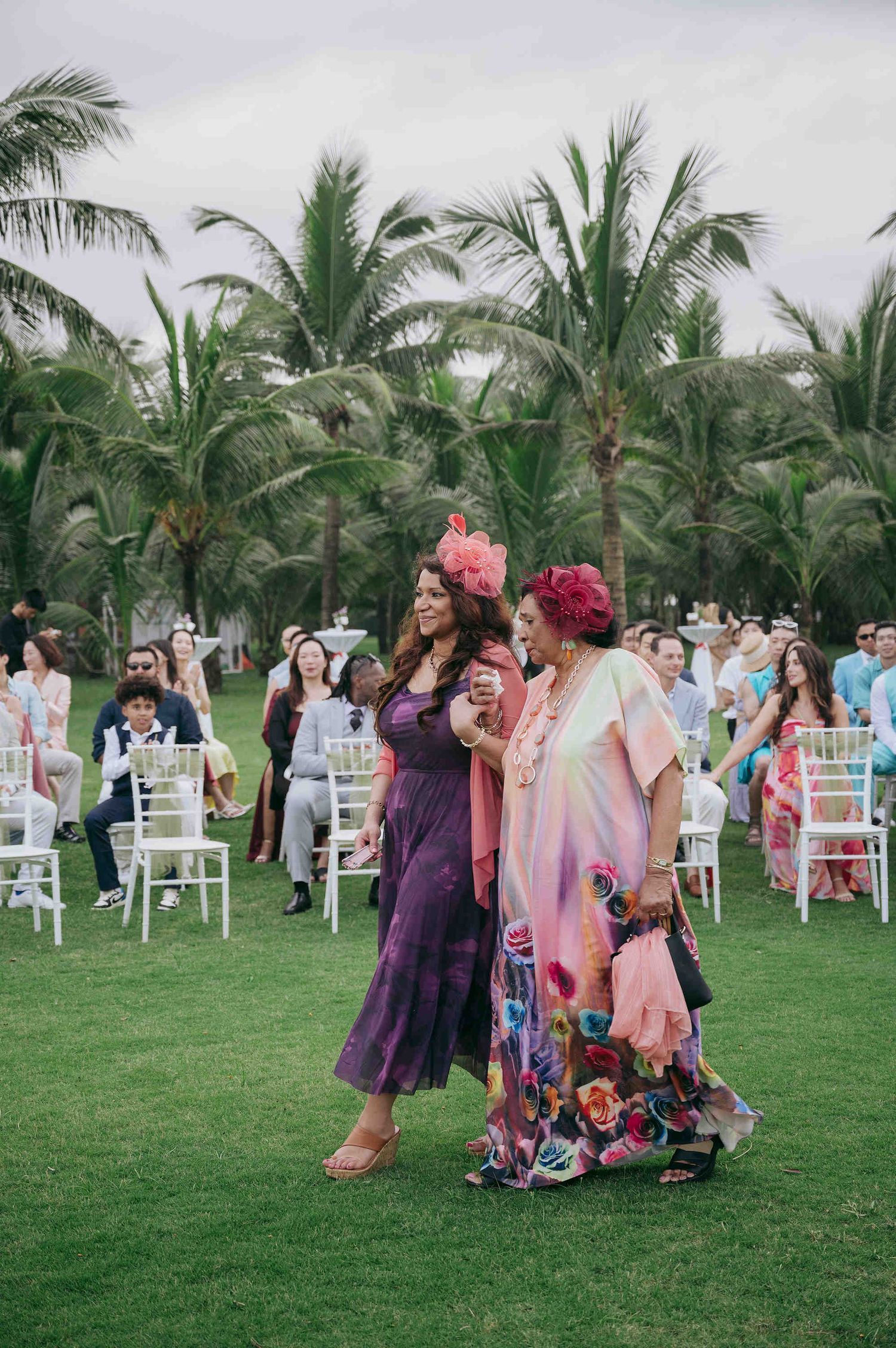 Guests in colorful attire walk across a lush green lawn at an outdoor wedding ceremony with palm trees.