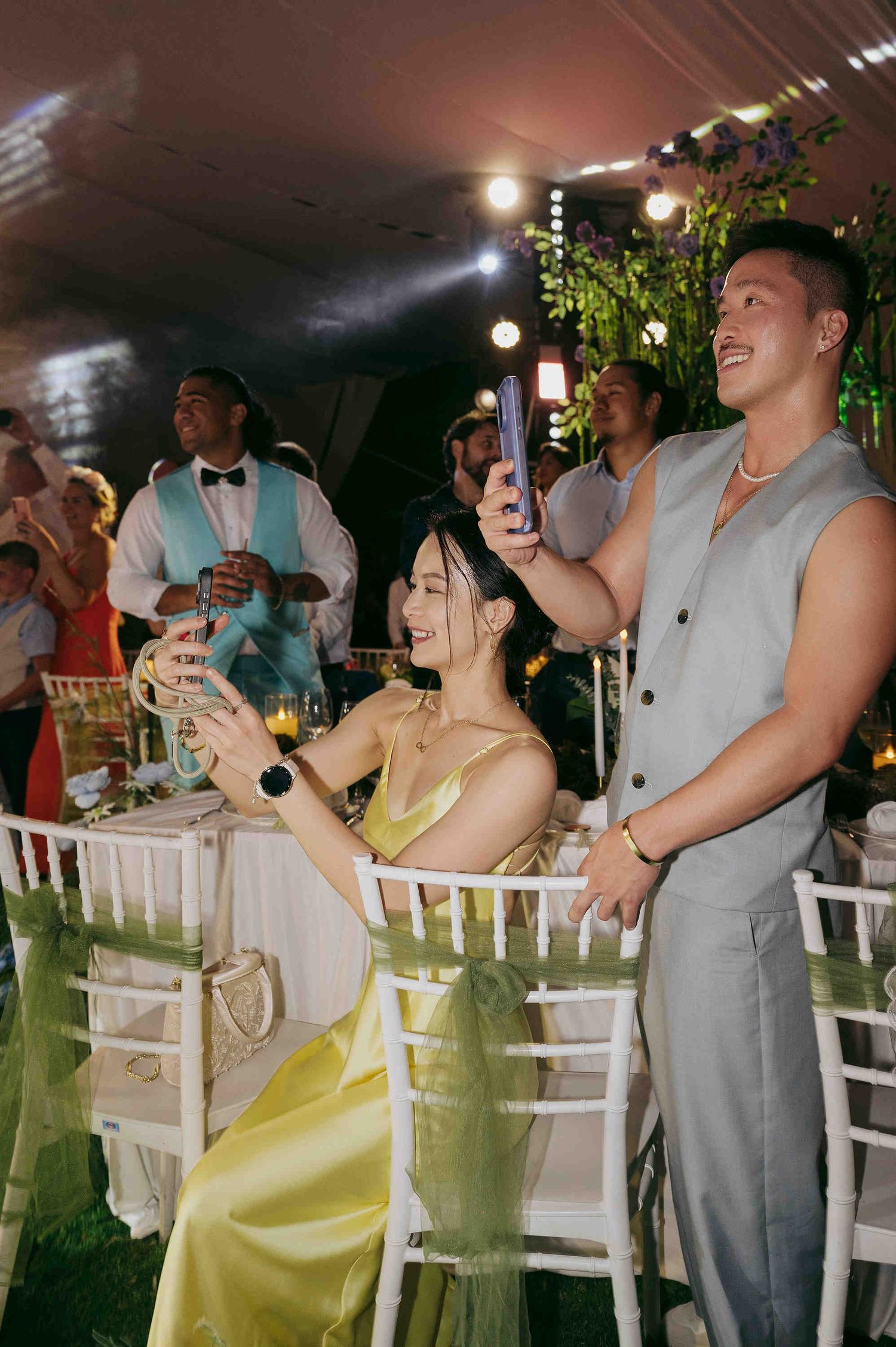 Guests in formal attire smile and take photos at an elegant outdoor wedding reception with string lights.