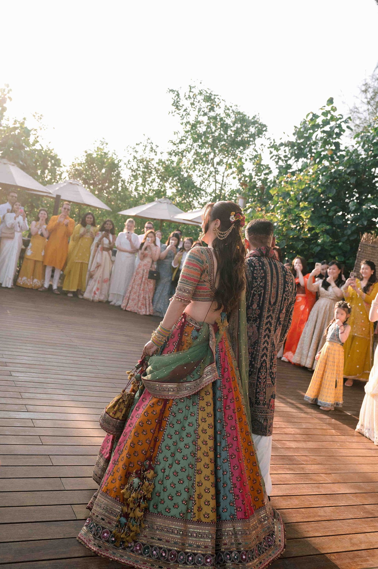 Bride in colorful lehenga walks with groom at outdoor Indian wedding celebration with guests in traditional attire.