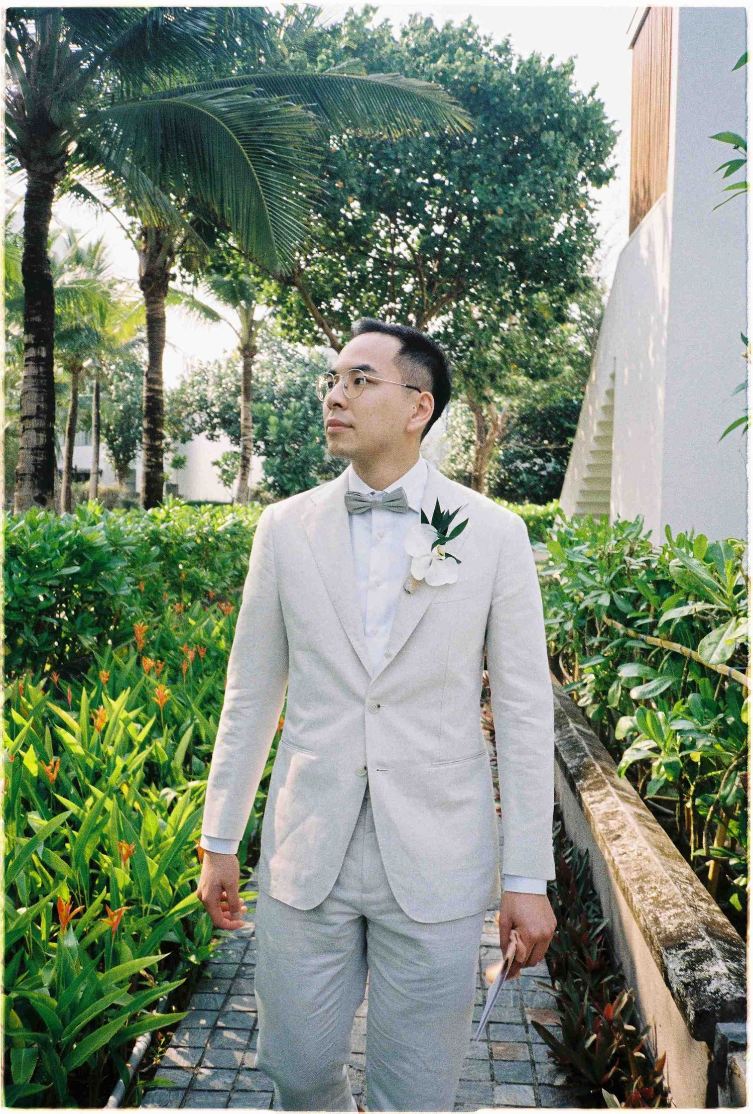 Groom in light gray linen suit with bow tie and boutonniere standing in lush tropical garden on wedding day.
