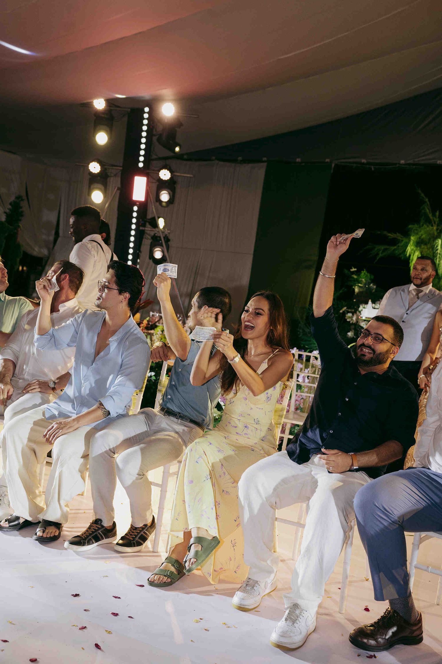 Guests cheer and raise phones at an elegant outdoor evening event, seated in white chairs under warm string lights.