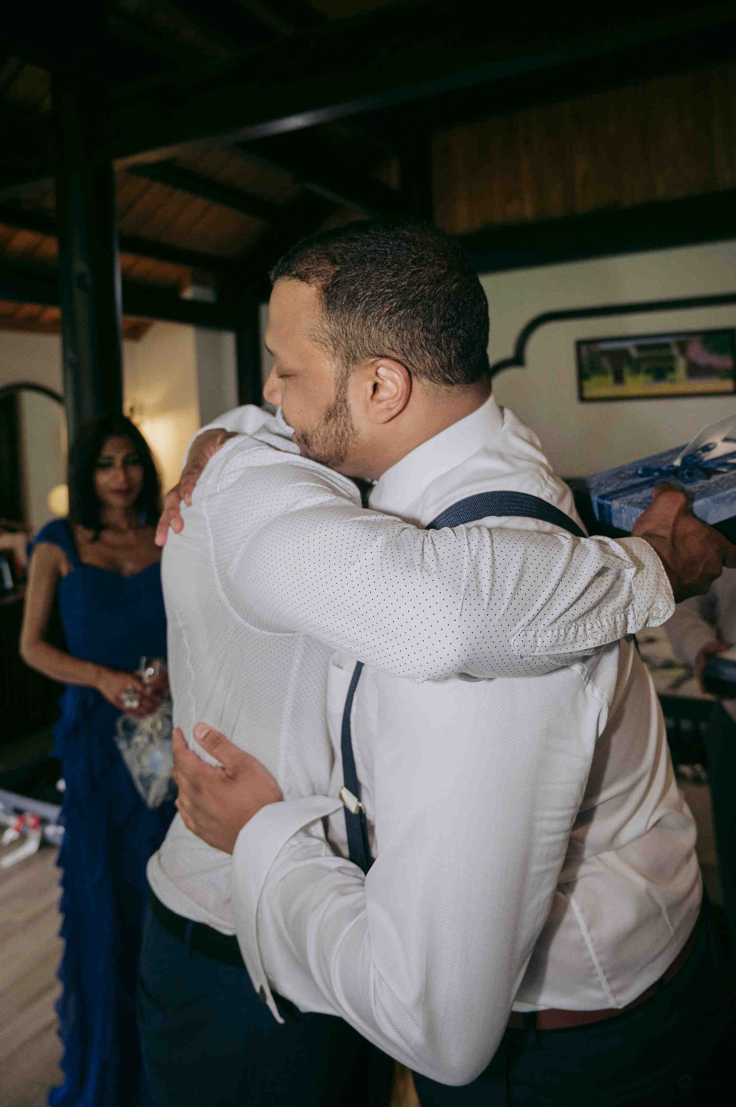 Two men in white dress shirts embrace warmly at a wedding celebration indoors.