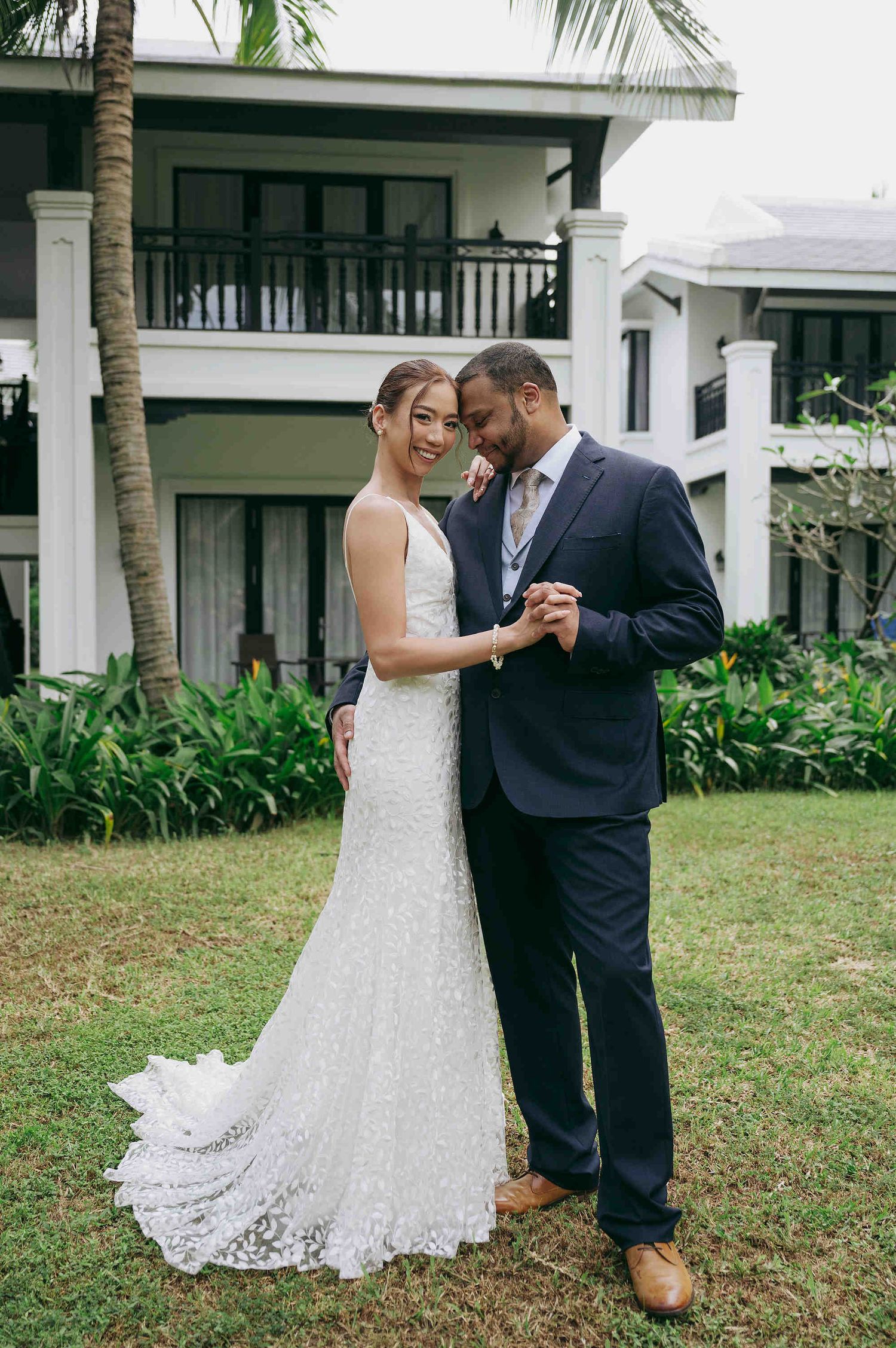 Bride in lace gown and groom in navy suit hold hands smiling in front of tropical villa on wedding day.