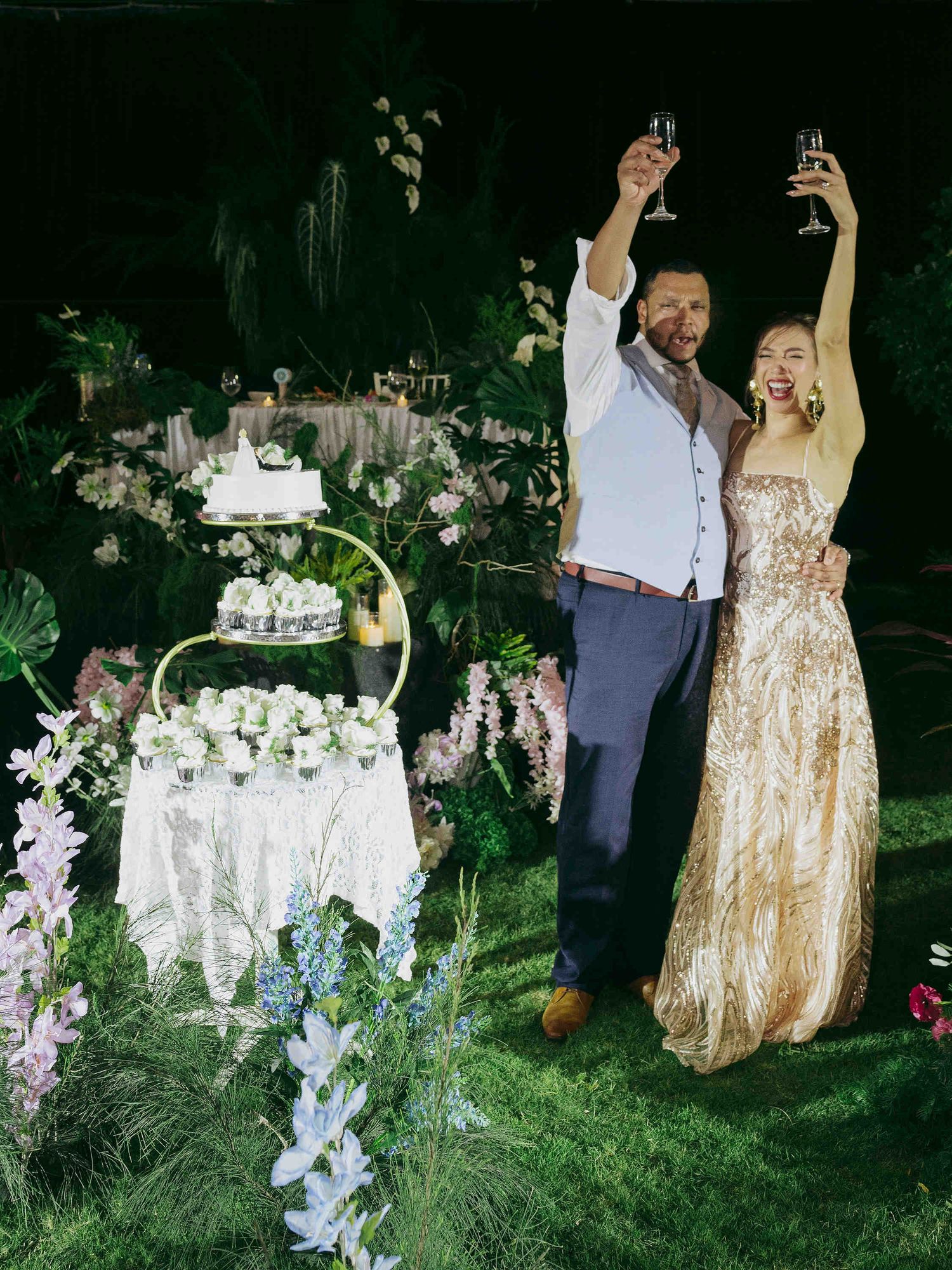 Couple in elegant attire toasting with champagne beside a floral wedding cake in a lush garden setting.