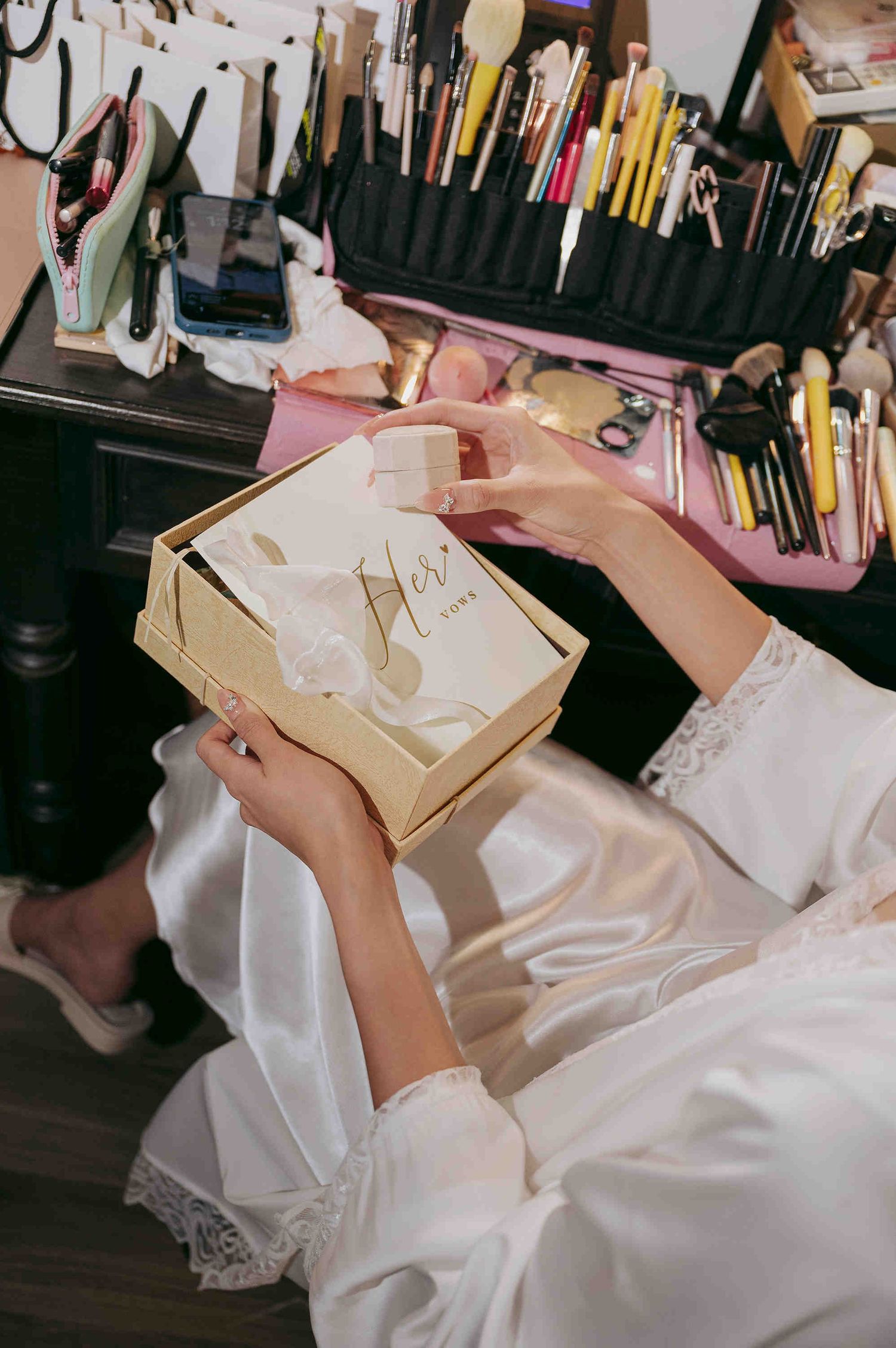 Bride in white satin robe opening a gold gift box at a makeup vanity filled with professional brushes and beauty tools.