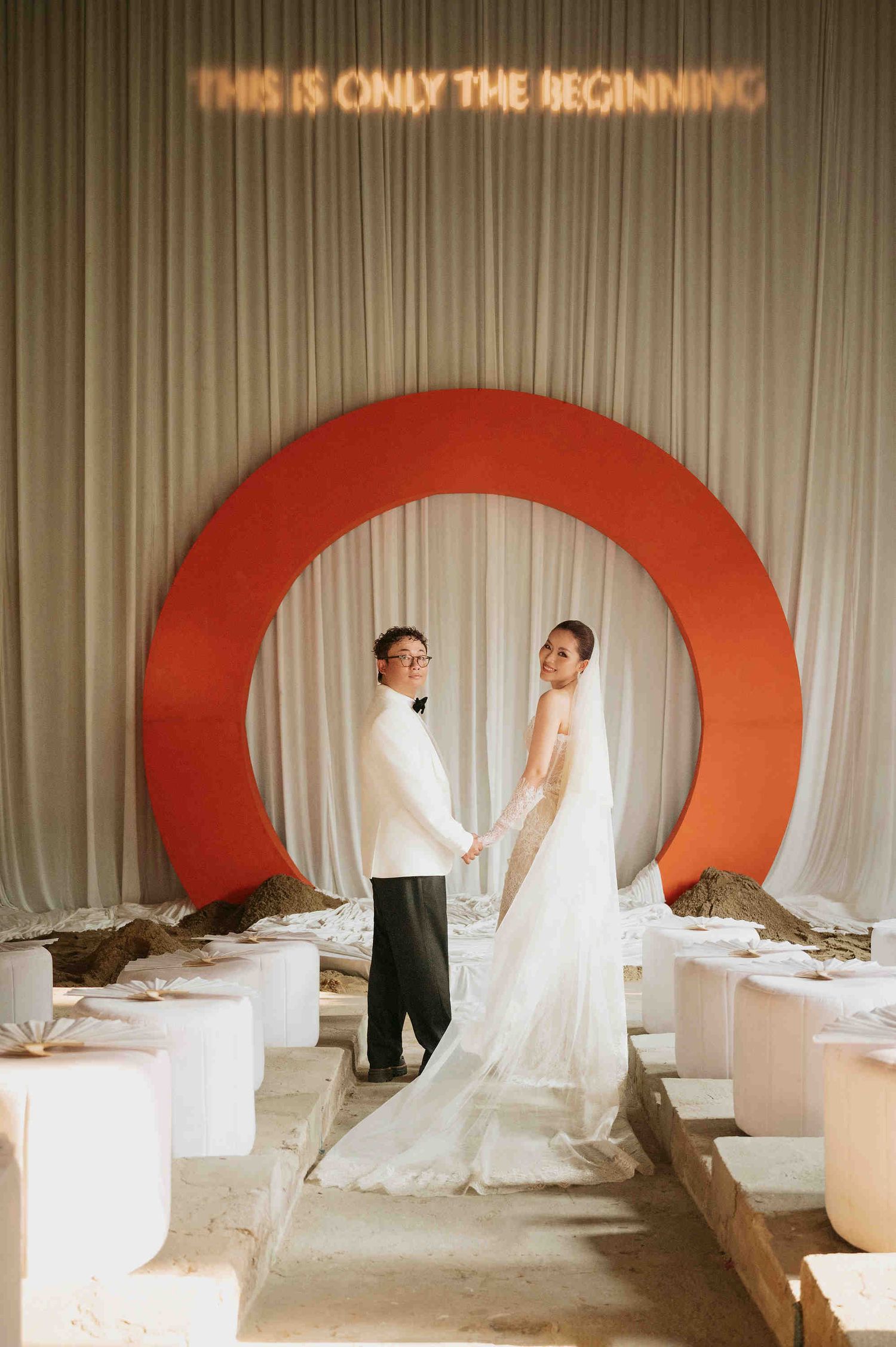 Couple poses in front of red circular arch with 'This Is Only The Beginning' neon sign at elegant wedding venue.