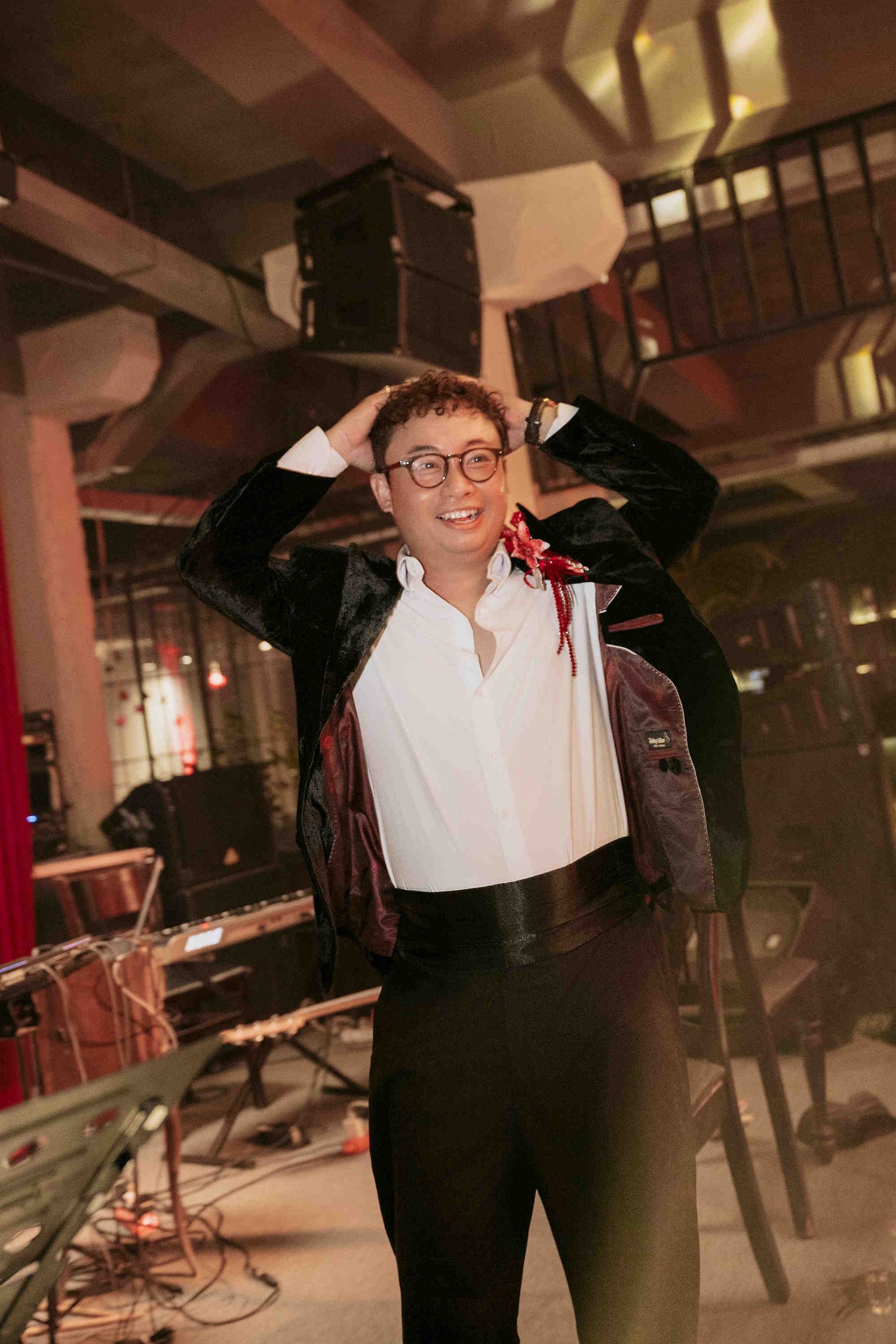 Smiling person in glasses, white shirt, and black velvet jacket posing with hands on head at a music venue.