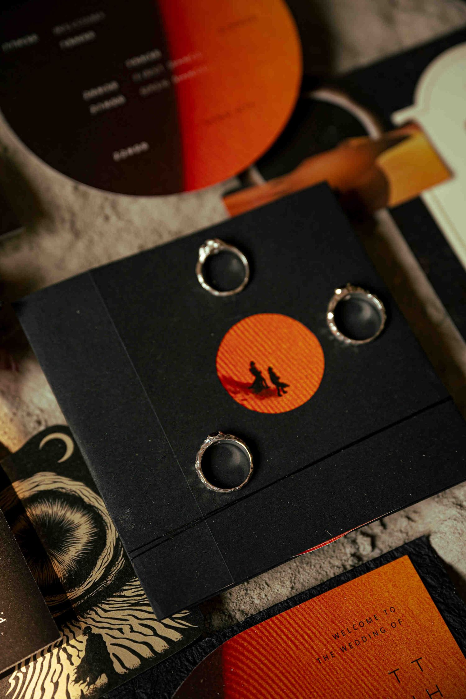 Black wedding ring box with silver rings on orange circle design, surrounded by elegant dark wedding stationery.