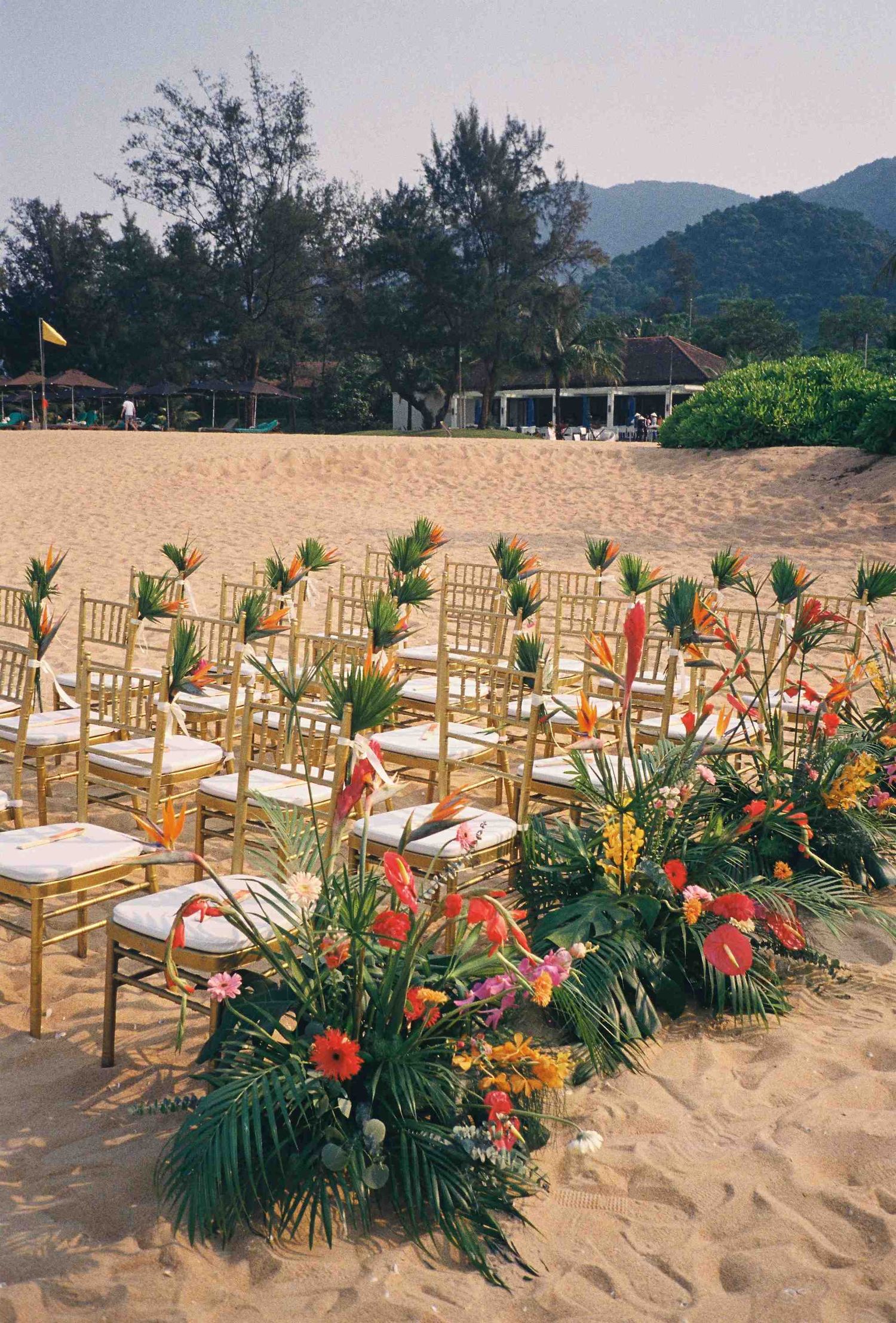 Lang Co wedding: beach wedding setup with gold chairs and tropical floral arrangements on sandy shore, by AnhVuWedding.