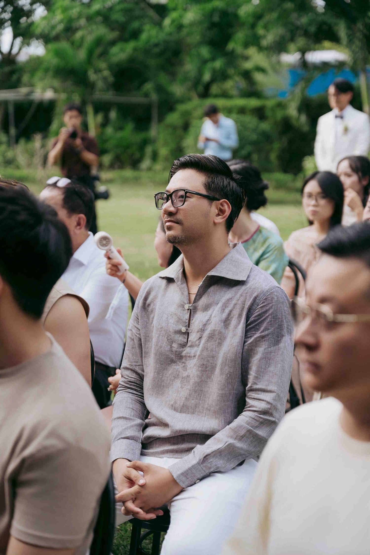 Man in glasses and linen shirt seated outdoors at a wedding ceremony, looking up attentively.