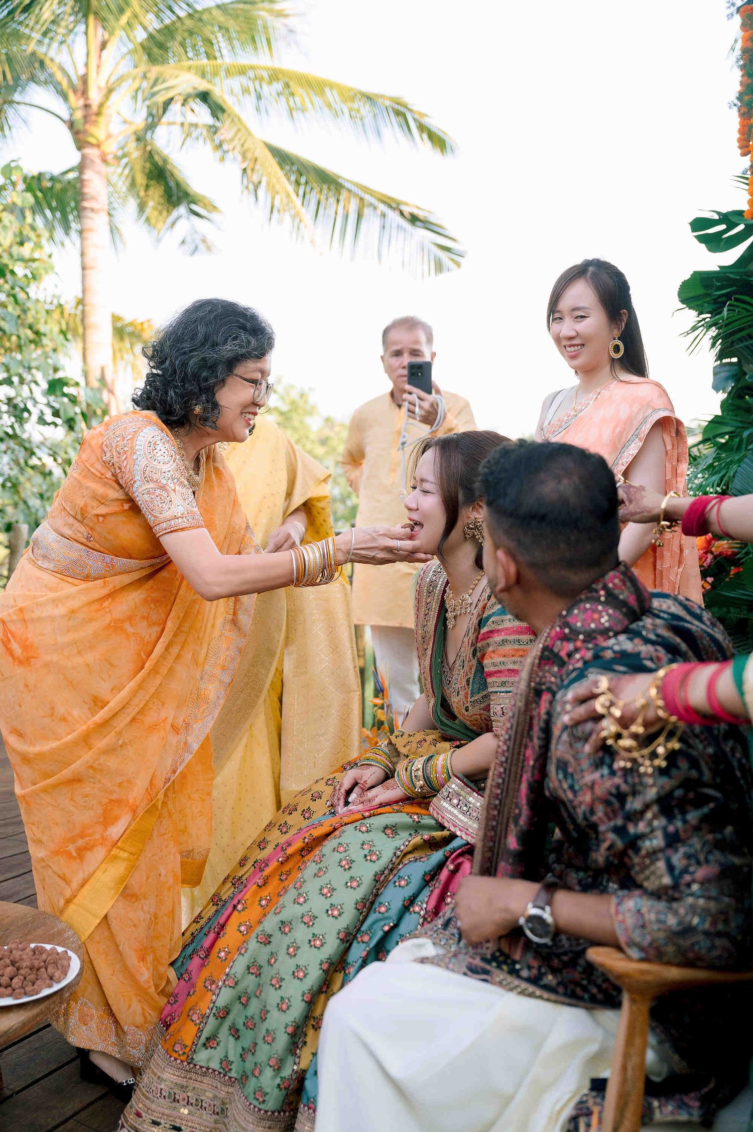 Indian wedding ceremony with guests in colorful traditional attire, bride receiving blessings outdoors with palm trees.