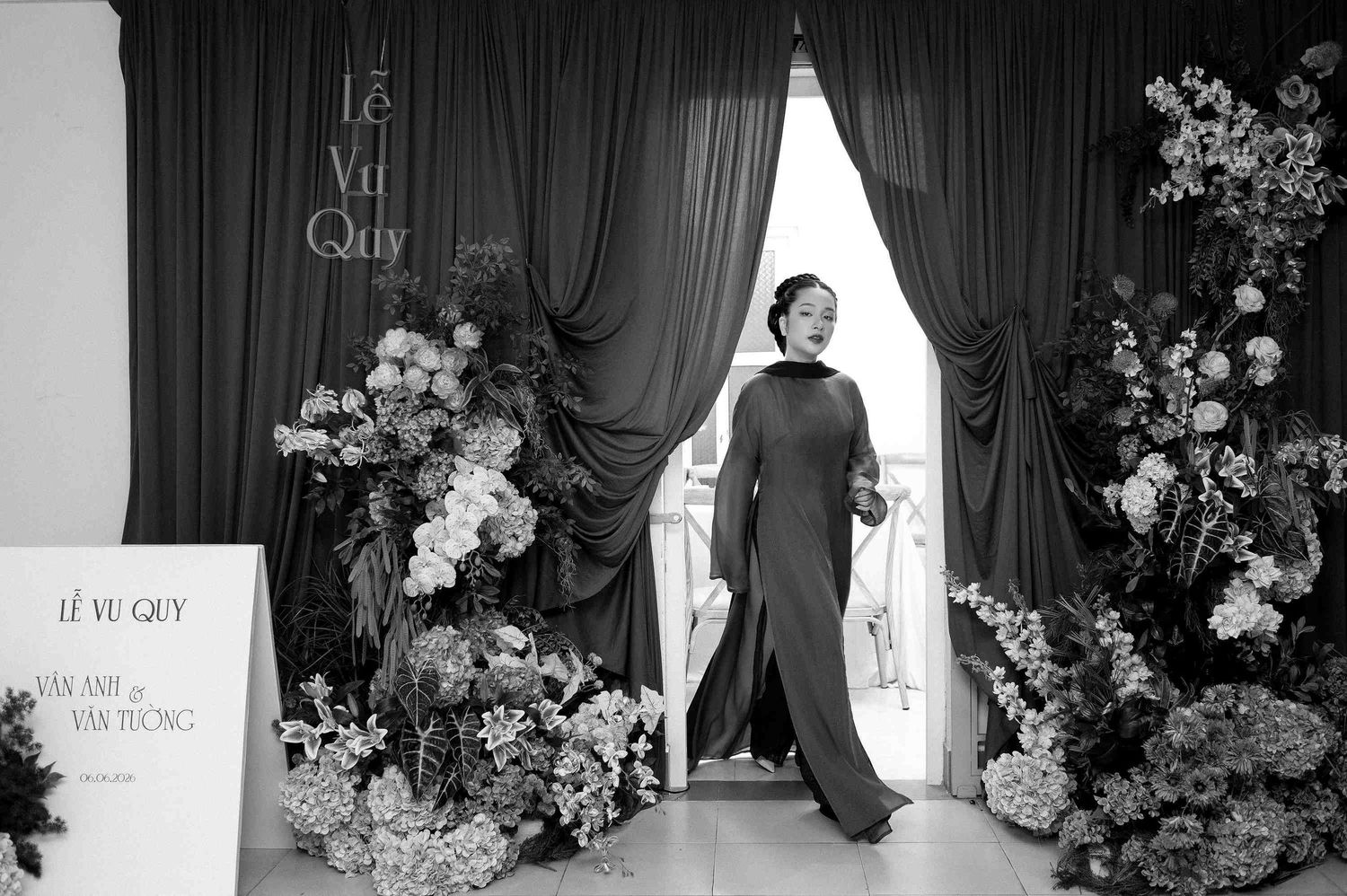 Black and white photo of a woman in an ao dai gown at a Vietnamese wedding ceremony with floral arrangements.