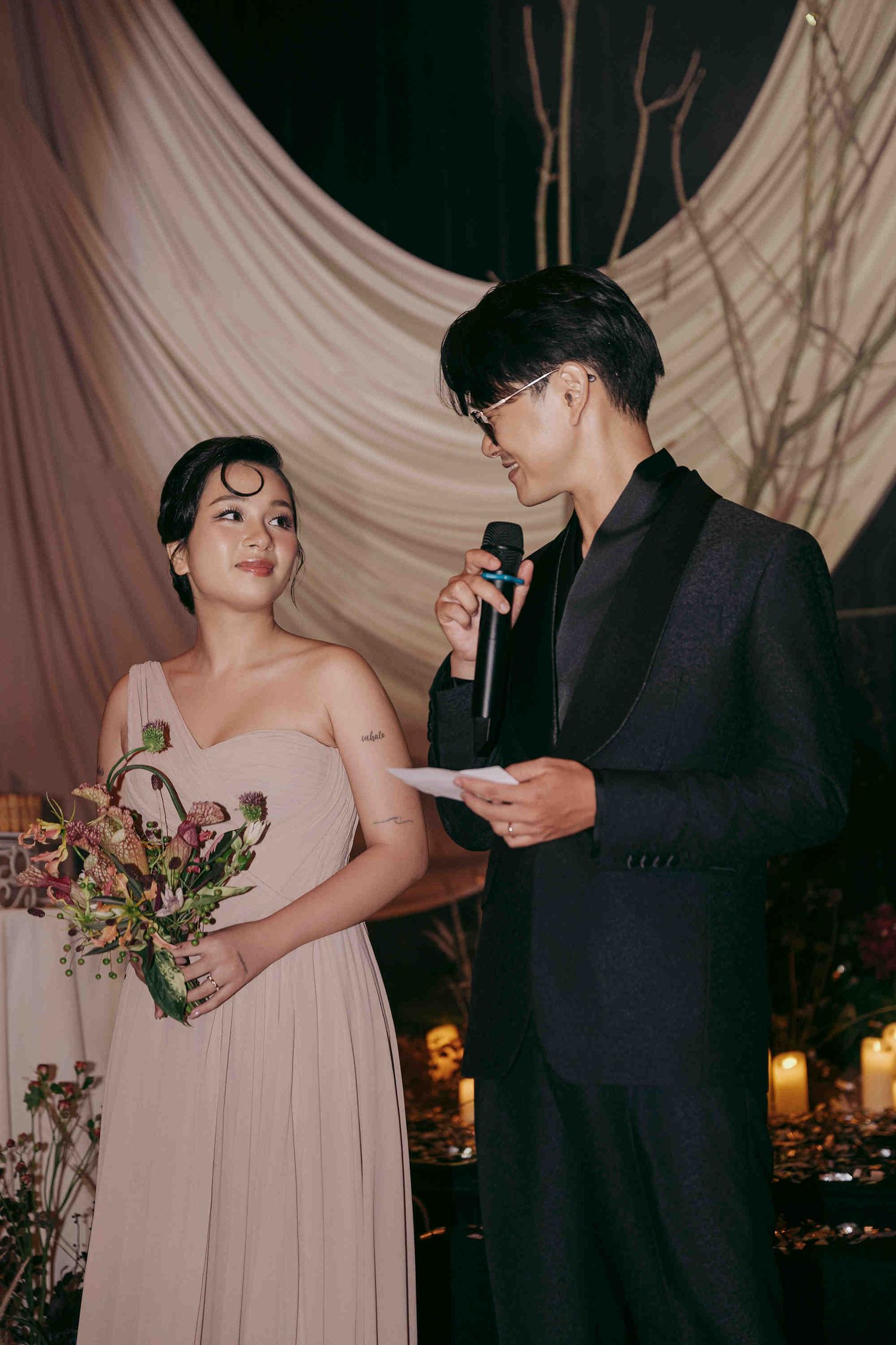 Couple in formal attire at wedding ceremony, groom reading vows into microphone while bride holds bouquet.