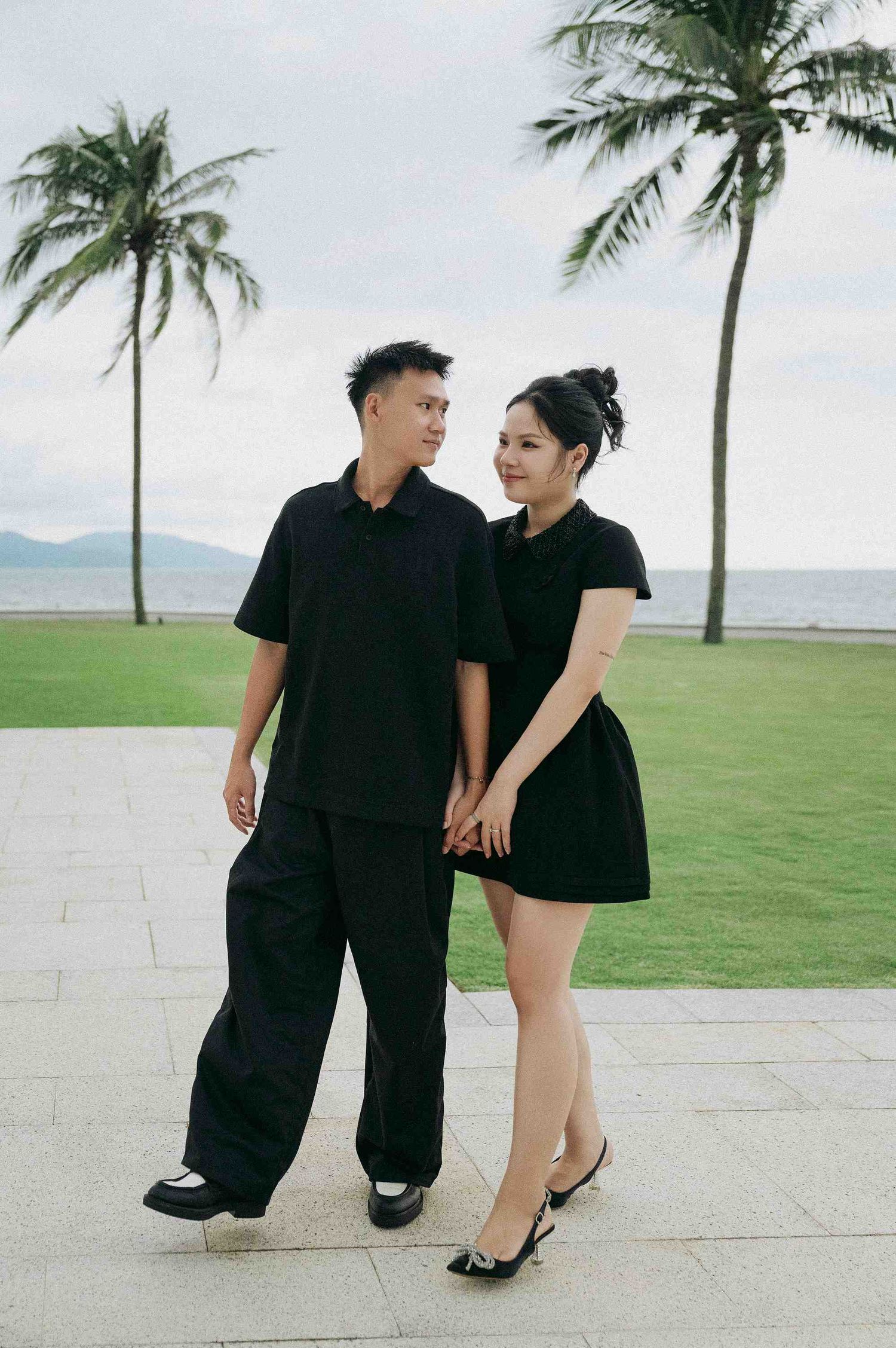 Da Nang family: two people in black attire stroll together on a scenic oceanfront walkway, by AnhVuWedding.