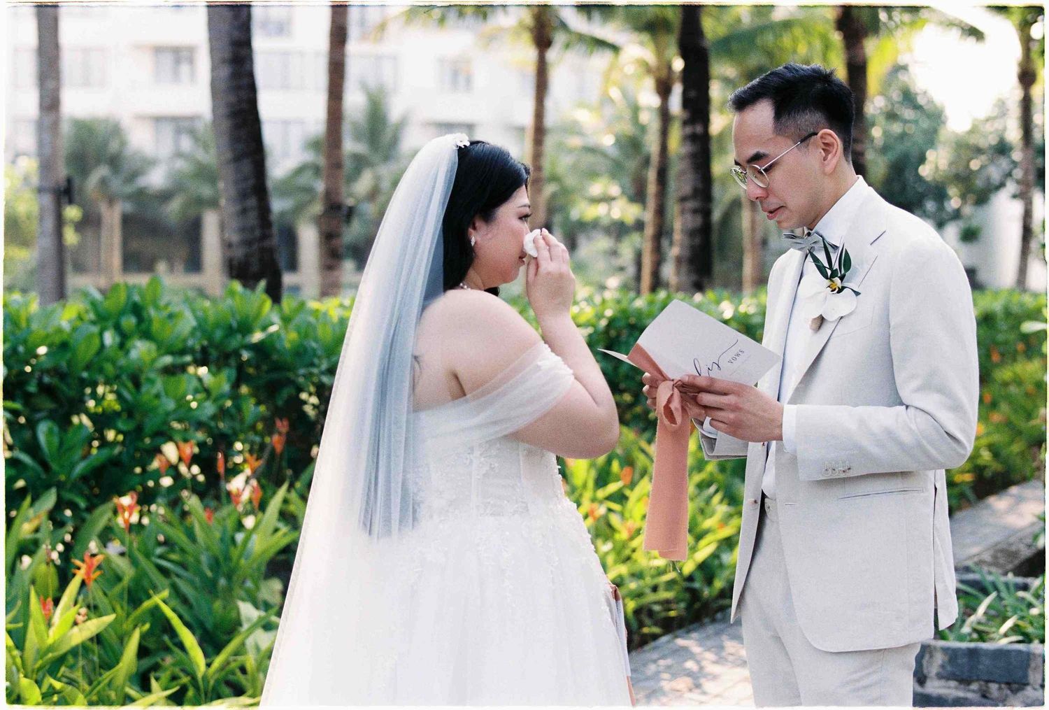 Bride wiping tears as groom reads wedding vows during outdoor ceremony, surrounded by tropical greenery.