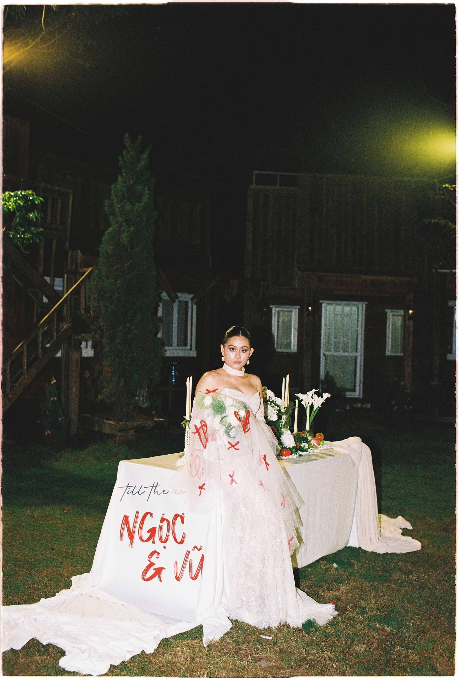 Saigon pre-wedding: bride in pink floral gown poses outdoors at night next to a decorated white table with a name sign, by AnhVuWedding.