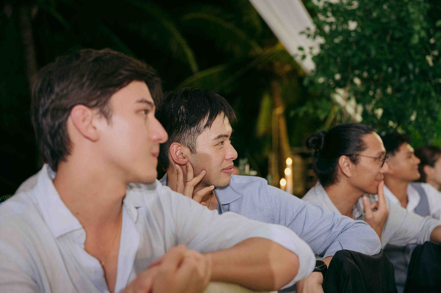 Four men in formal attire seated outdoors at an evening event, attentively watching a wedding ceremony.