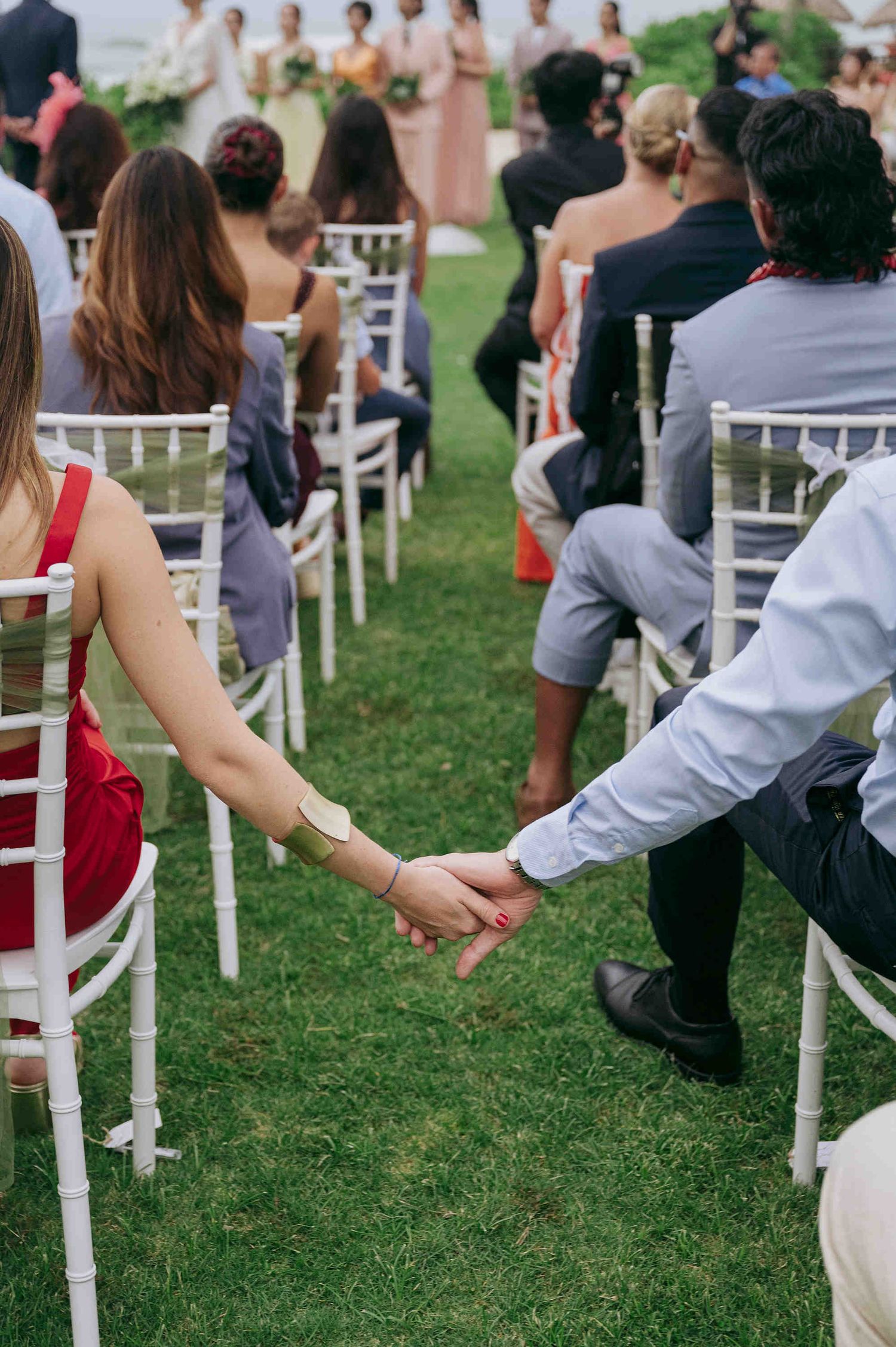 Wedding guests hold hands while seated in white chairs at an elegant outdoor garden ceremony.