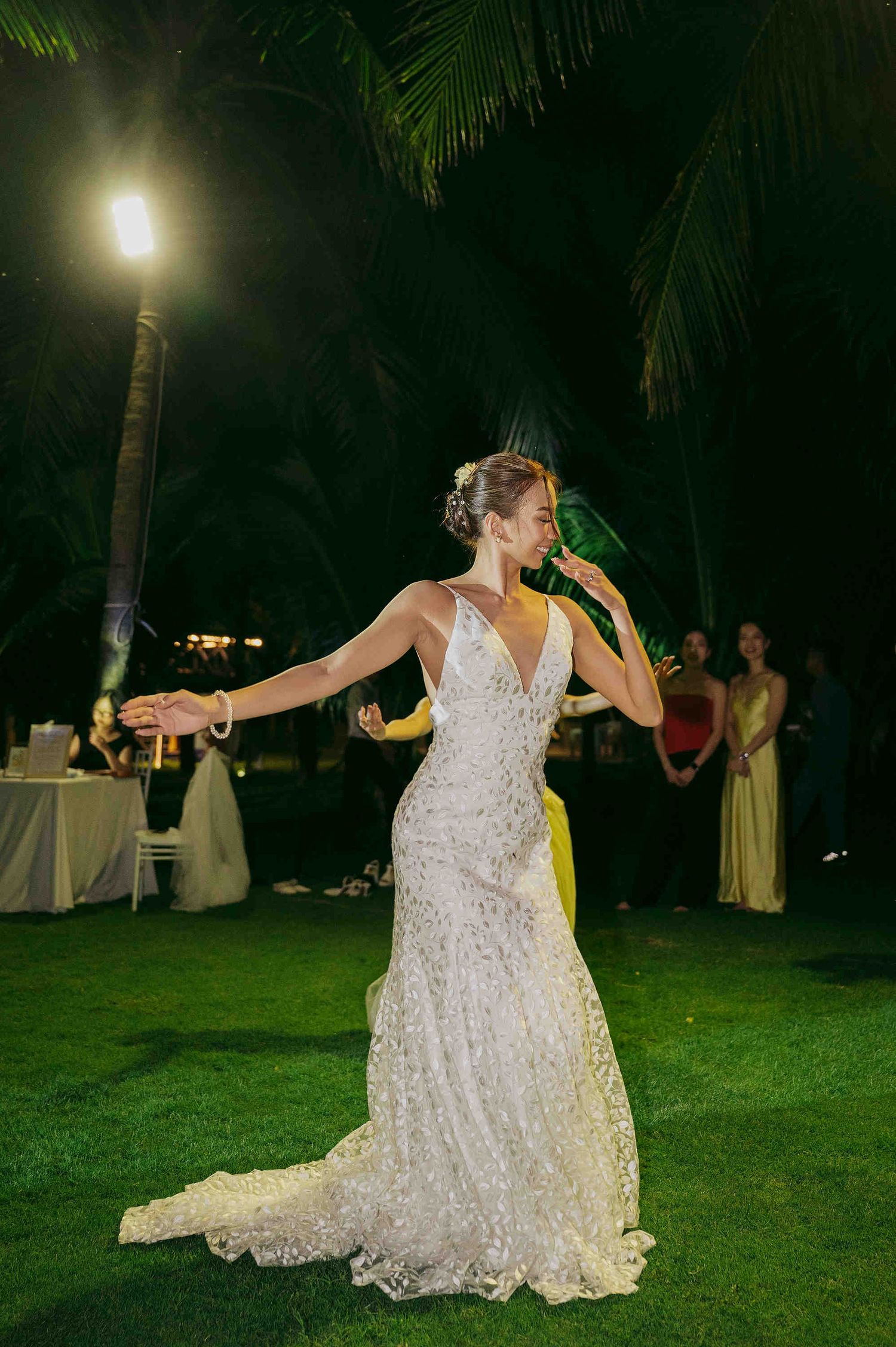 Bride in lace wedding gown dancing outdoors at night under palm trees at tropical wedding reception.