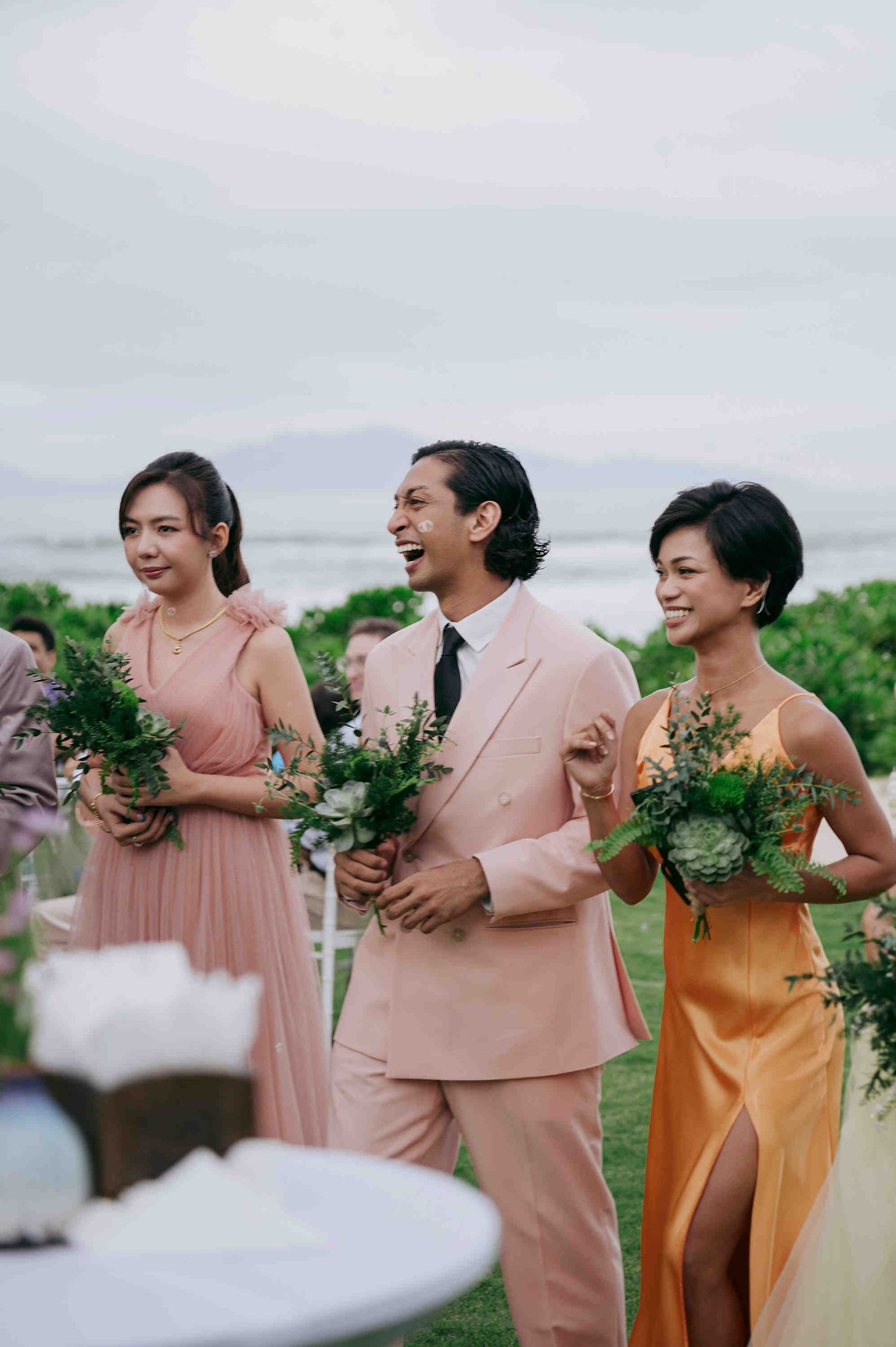Three wedding guests in pink and yellow attire holding bouquets at an outdoor ceremony.