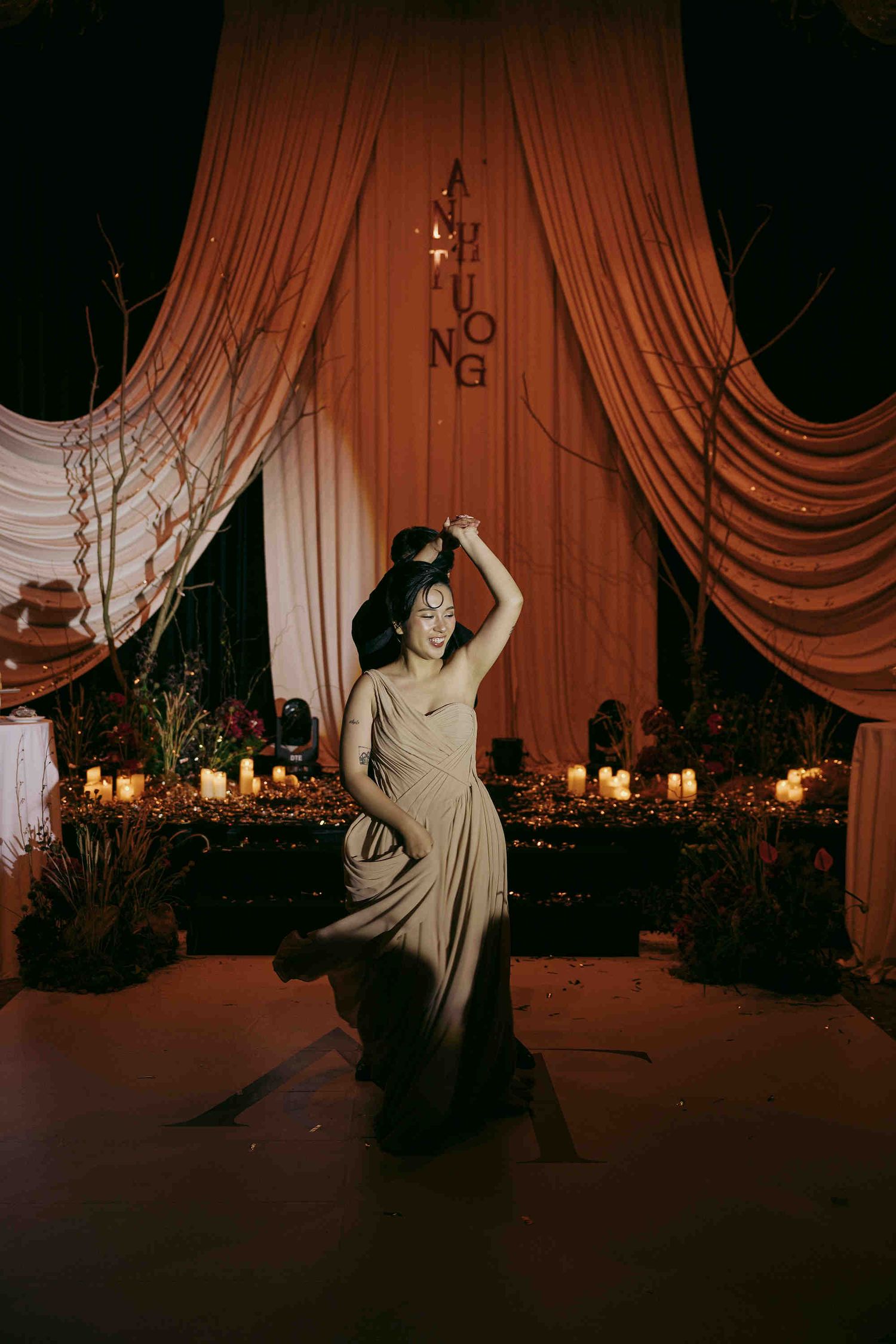Woman in elegant gray gown dancing gracefully before candlelit table with orange draped curtains backdrop.