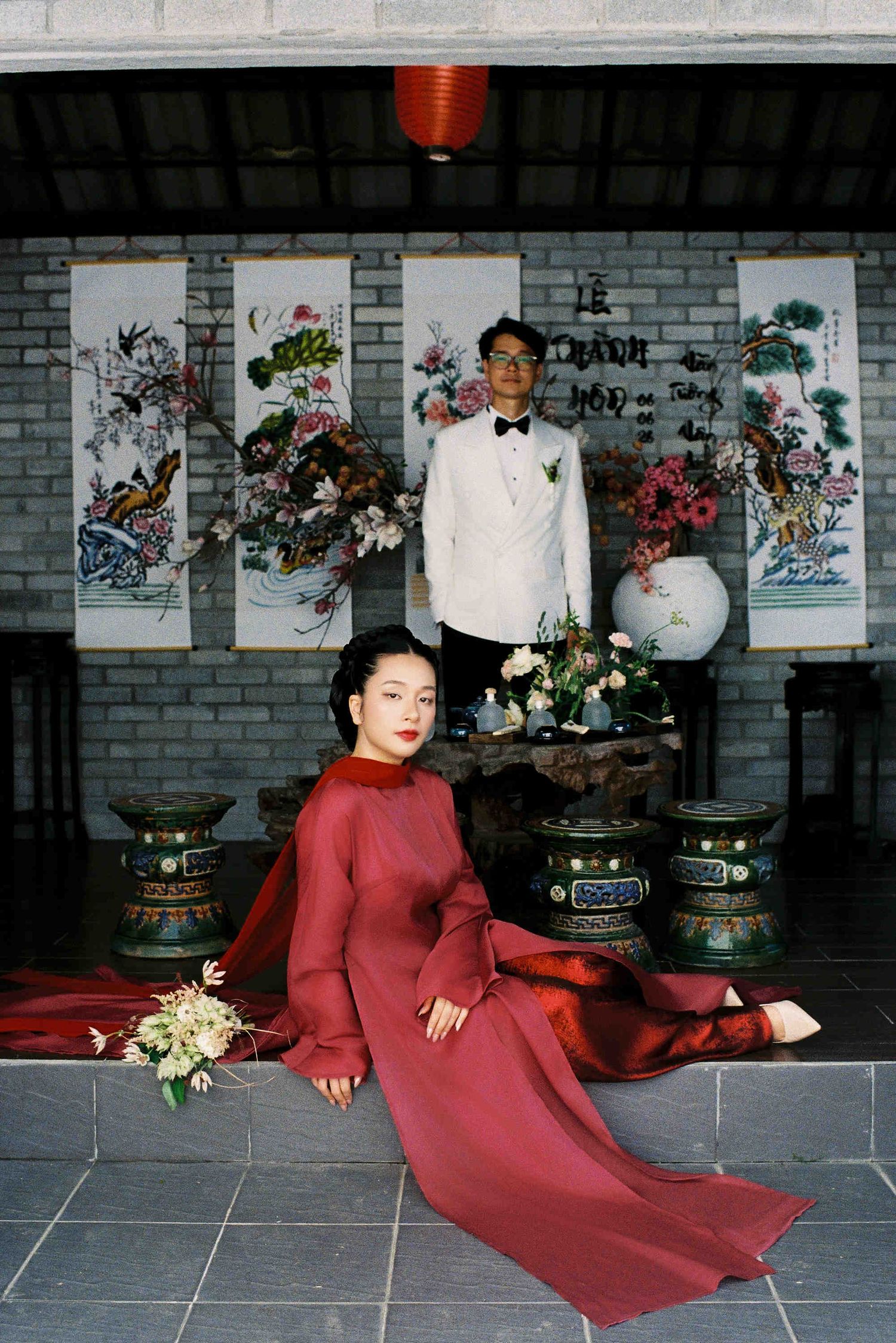 Vietnamese couple in traditional ao dai wedding attire pose in ornate temple setting with floral scrolls and red lantern.