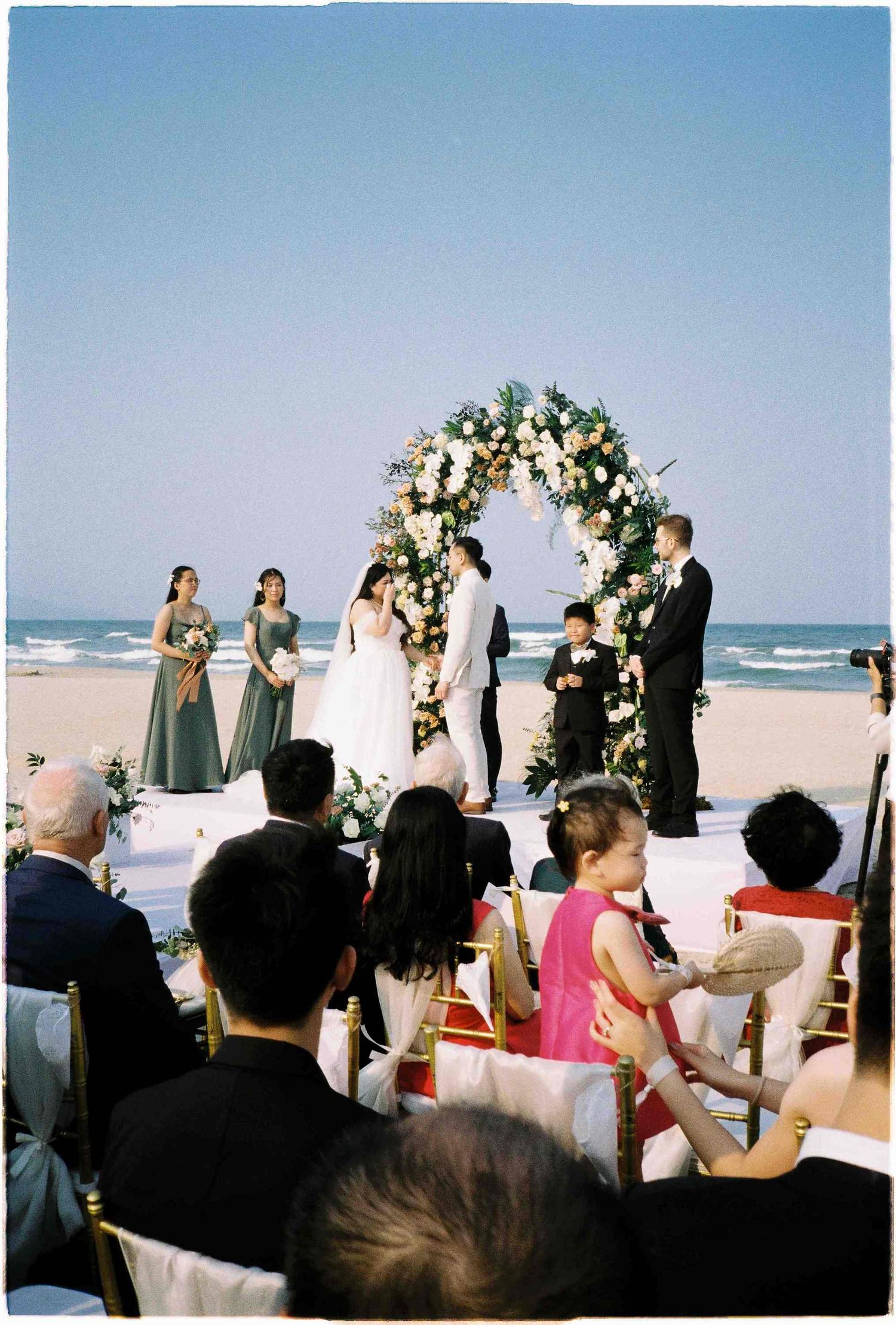 Beach wedding ceremony with floral arch, bride and groom, bridesmaids in sage green, and seated guests by the ocean.