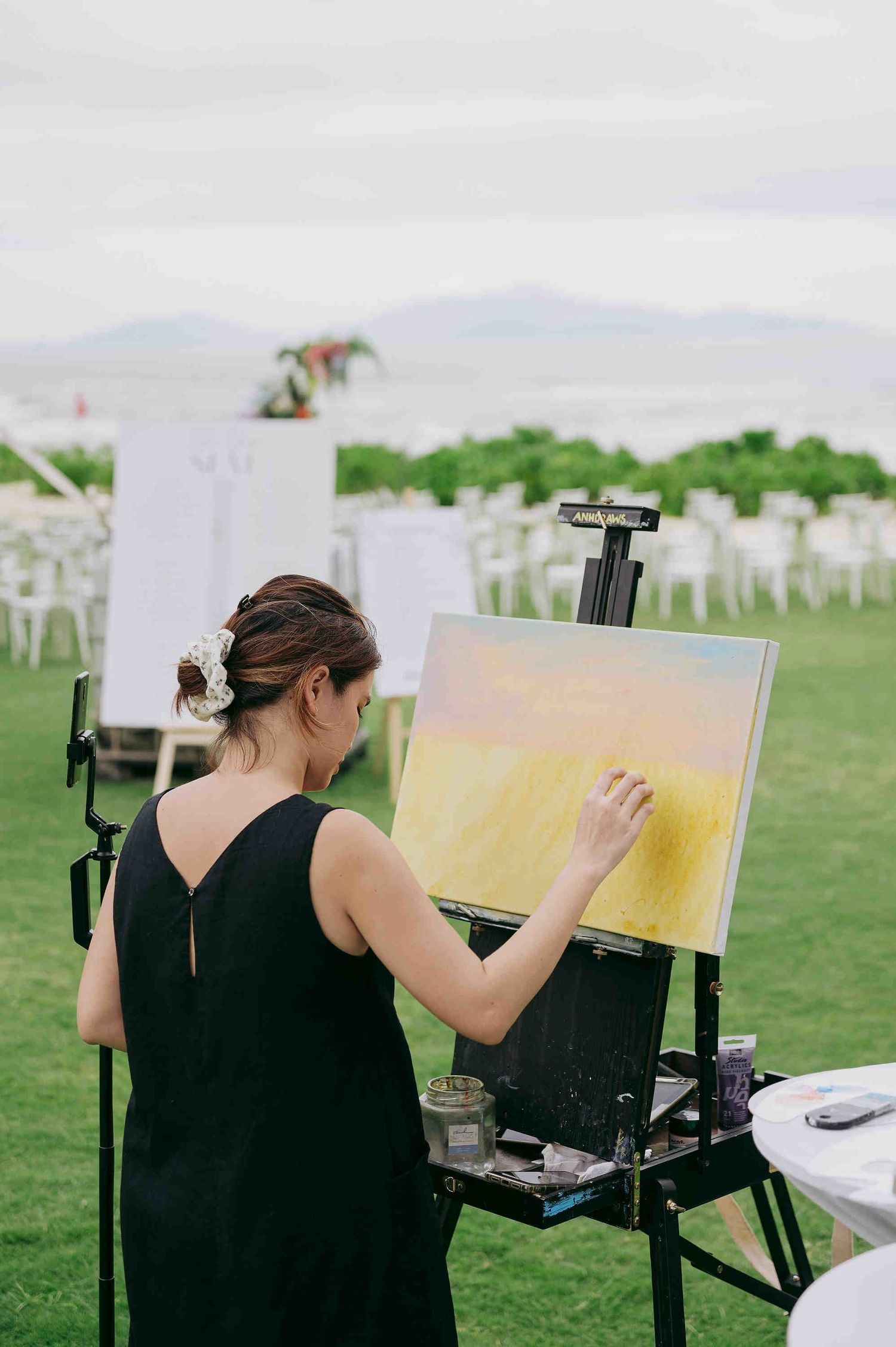 Woman in black dress painting on easel at outdoor wedding venue with white chairs and mountain backdrop.