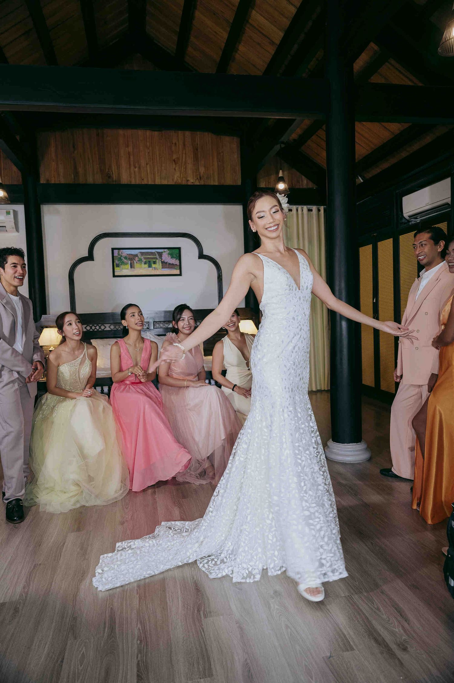Bride in white lace gown twirls before bridesmaids in colorful dresses at elegant indoor wedding venue.