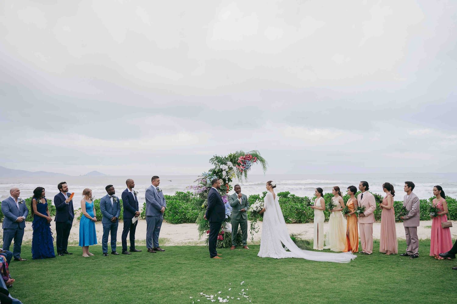 Outdoor wedding ceremony on green lawn with ocean backdrop, bridal party in colorful dresses, groomsmen in gray suits.