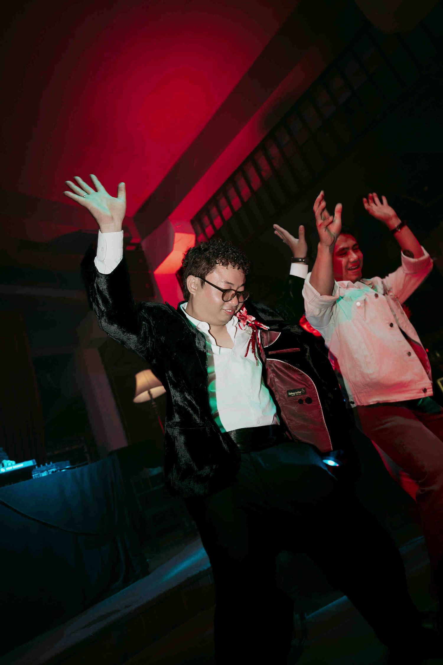 Two people dancing energetically at a party under red and green lights, arms raised in celebration.
