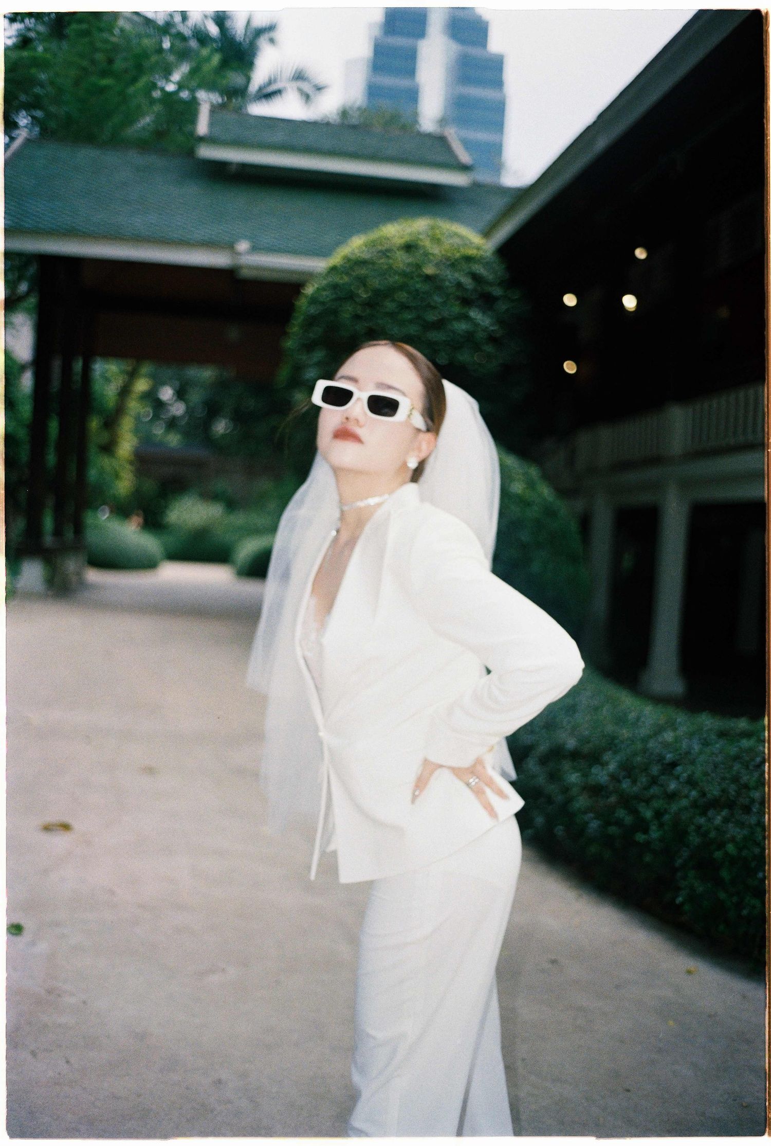 Saigon pre-wedding: figure in flowing white outfit and sunglasses poses dramatically in a garden setting, by AnhVuWedding.