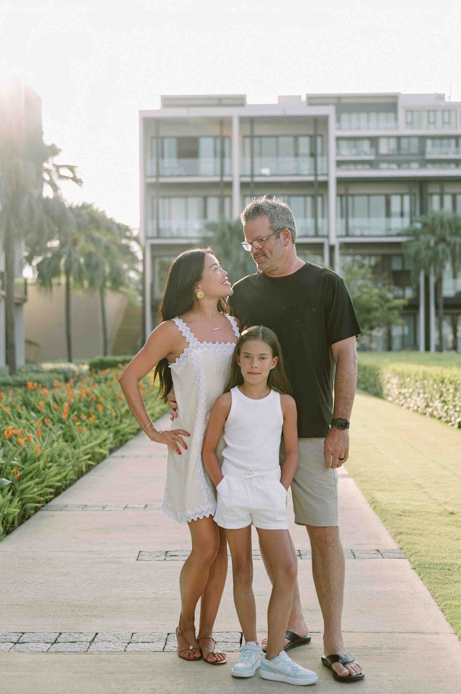 Da Nang family: family enjoying a sunset walk together on a beachfront pathway near modern condominiums and tropical, by AnhVuWedding.