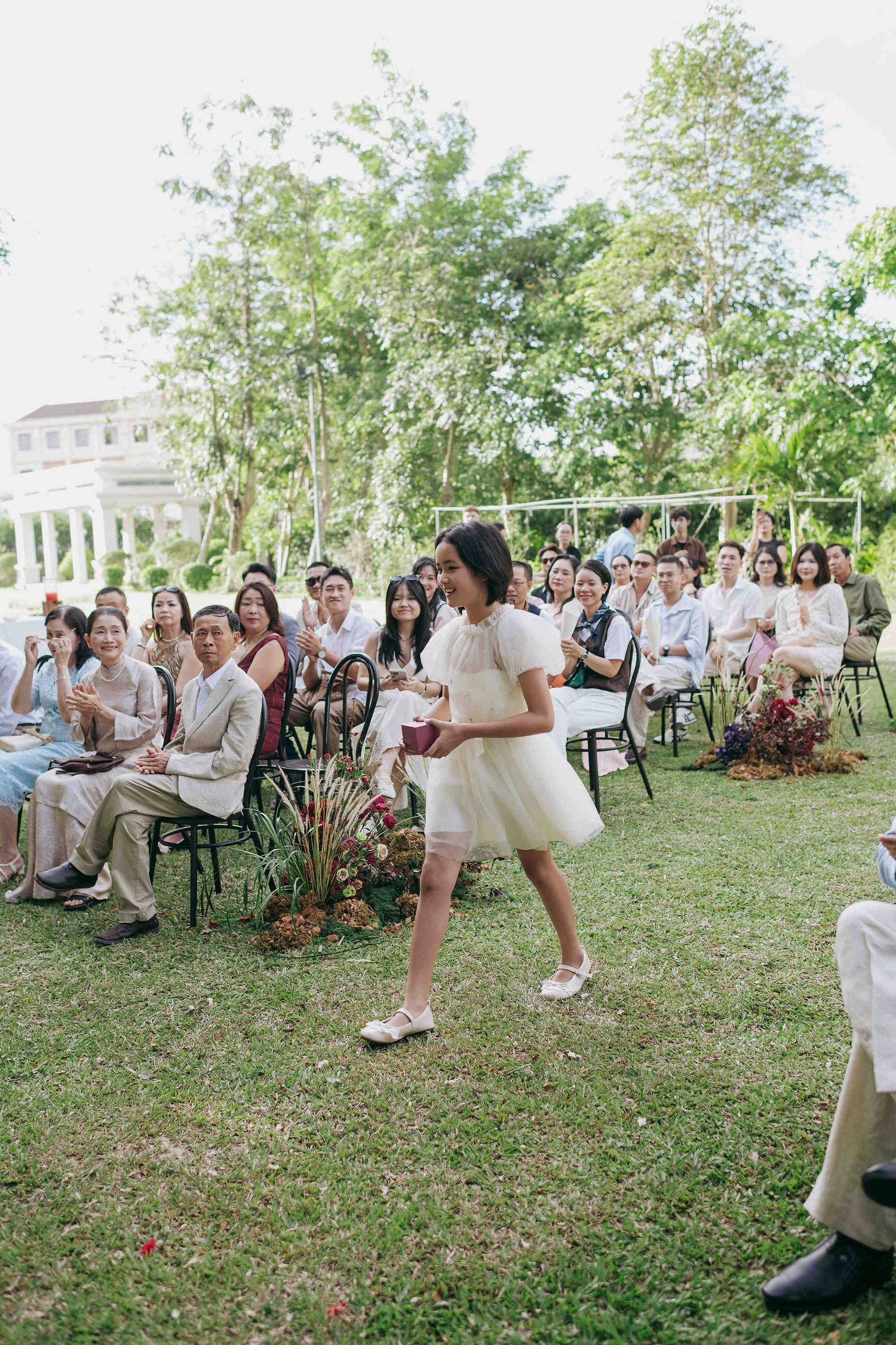 A flower girl in a white dress walks down the aisle at an outdoor wedding ceremony with guests seated on chairs.