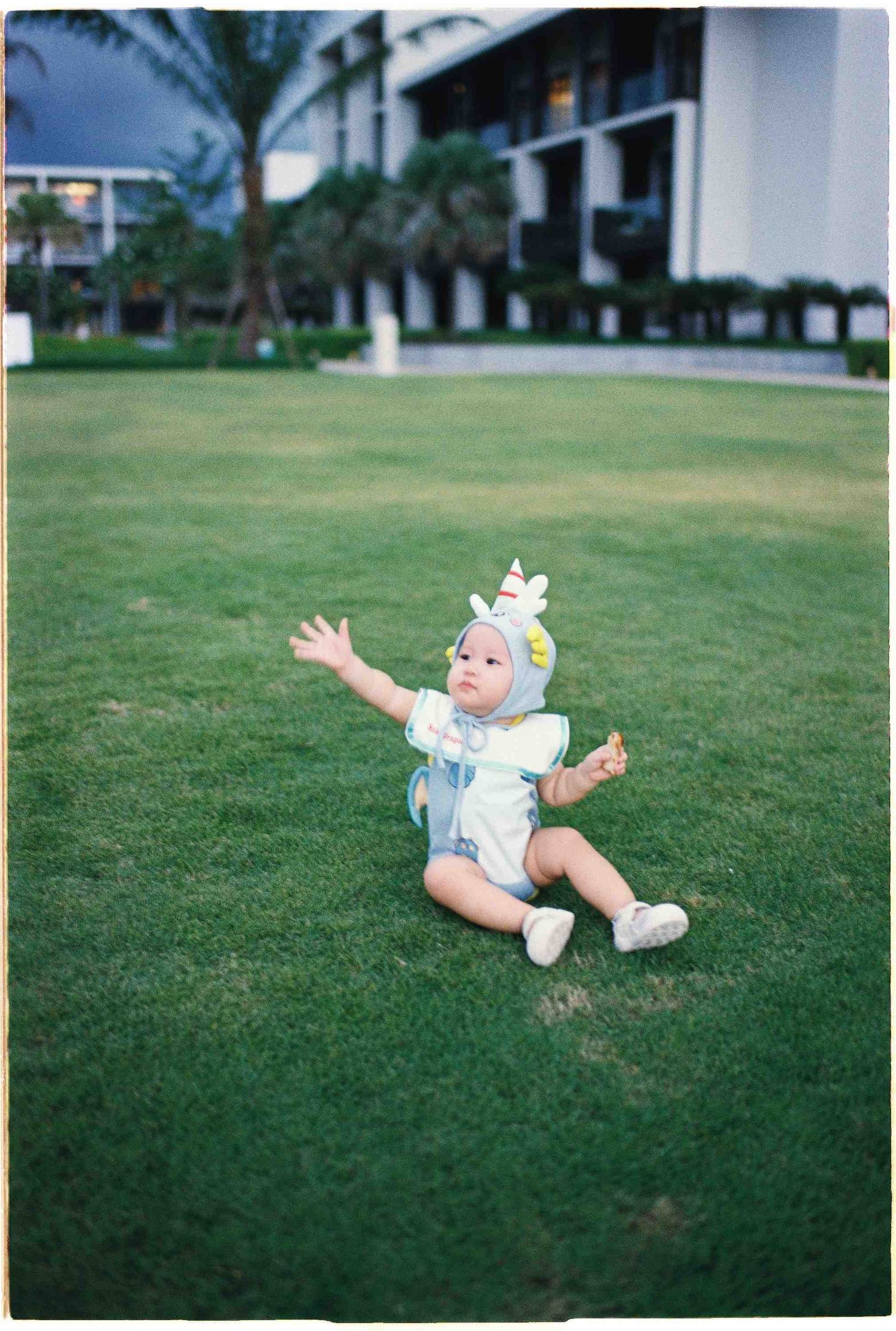 Da Nang family: baby wearing a cute costume sits on grass with arms raised at a resort property, by AnhVuWedding.