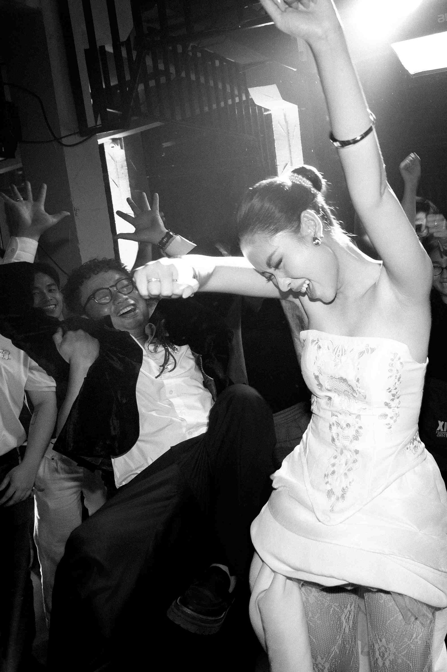 Black and white photo of a bride in white dress dancing joyfully with guests at a lively wedding reception.