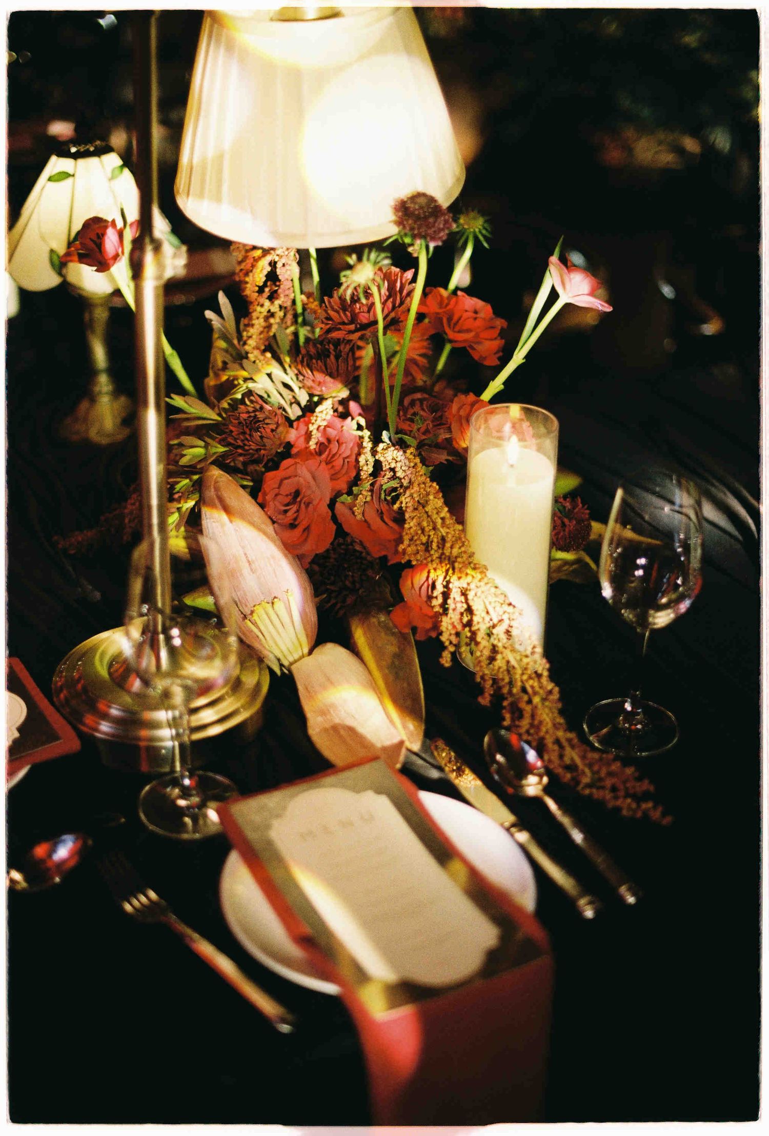 Elegant wedding table setting with warm lamp light, floral centerpiece, candles, wine glasses, and gold cutlery on dark linen.