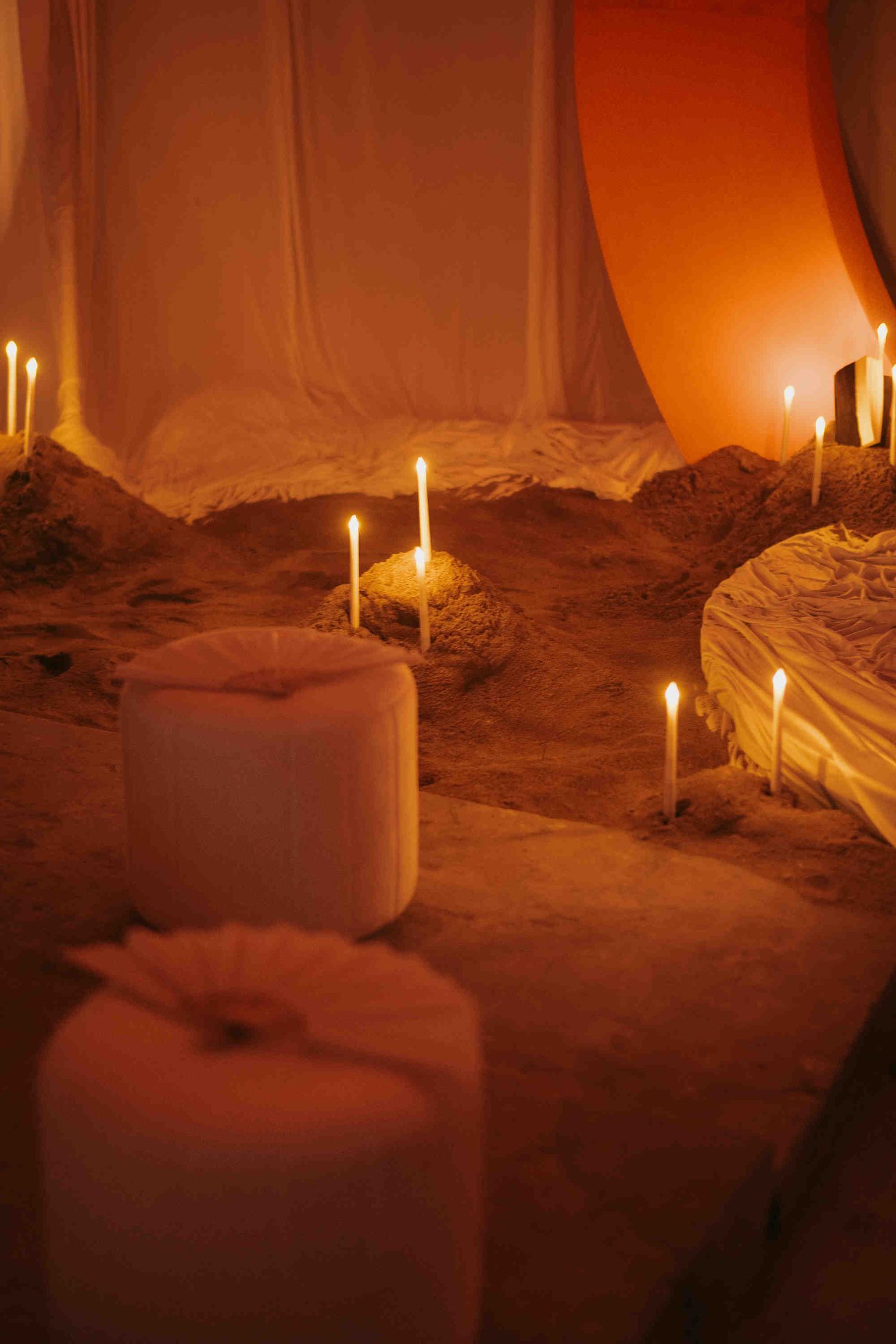 Romantic candlelit sandy setup with white draping and glowing candles in warm orange ambiance.