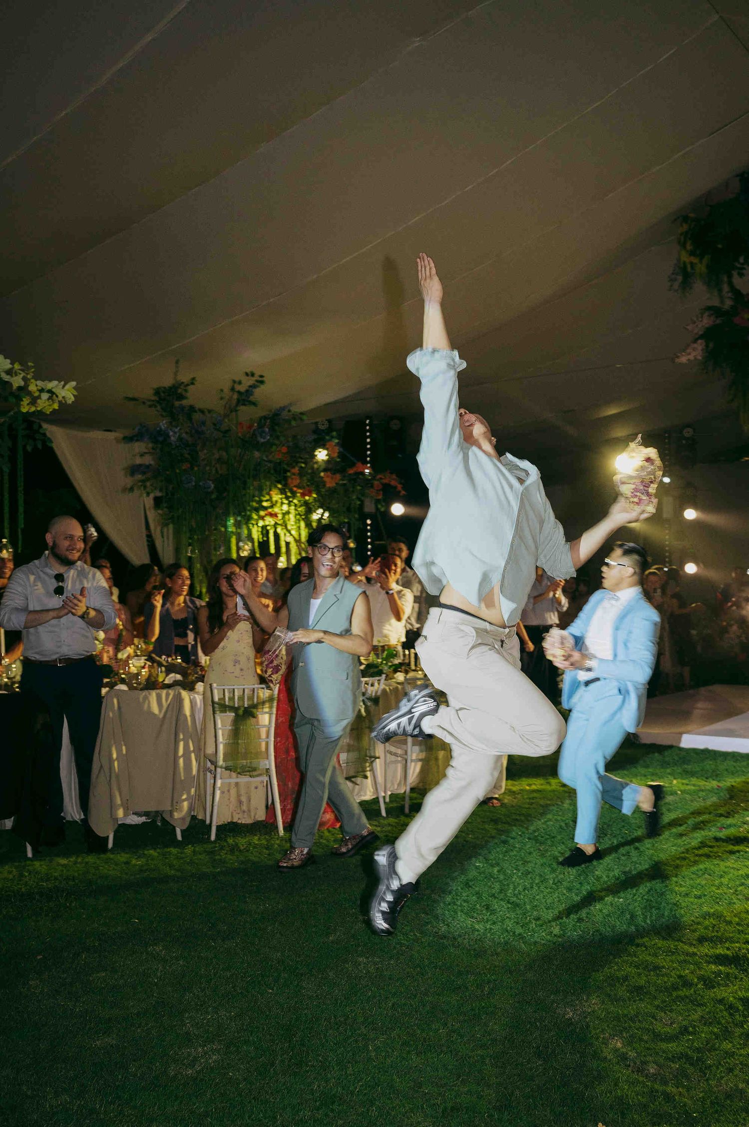 A groom leaps joyfully on a grass dance floor at an elegant tented wedding reception while guests celebrate around him.