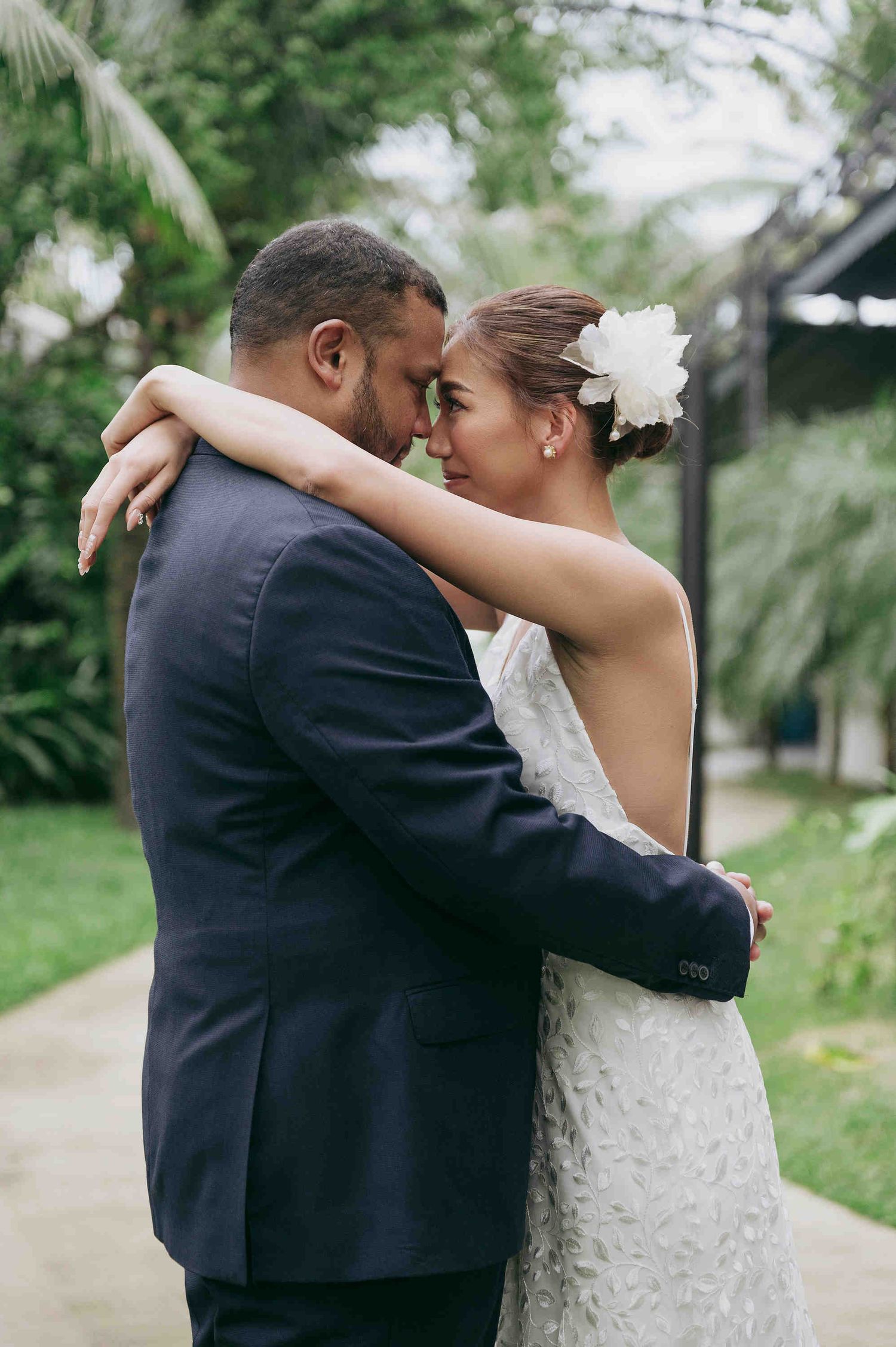 A bride and groom share an intimate moment, touching foreheads while embracing outdoors, surrounded by lush tropical greenery.