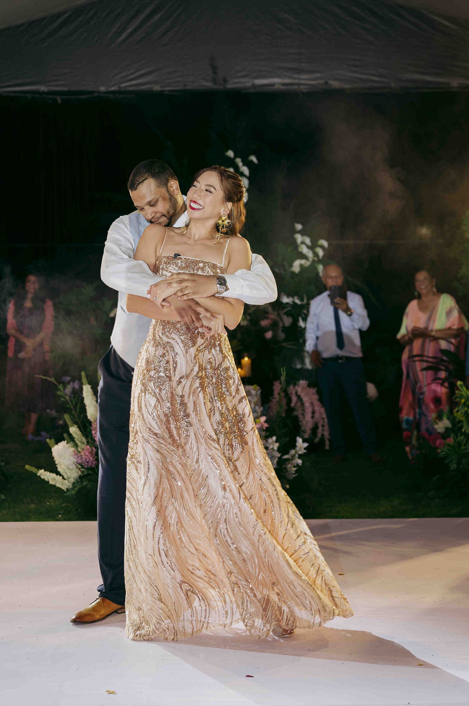 Couple sharing first dance at wedding, bride in gold sequin gown, groom in white shirt and navy pants.