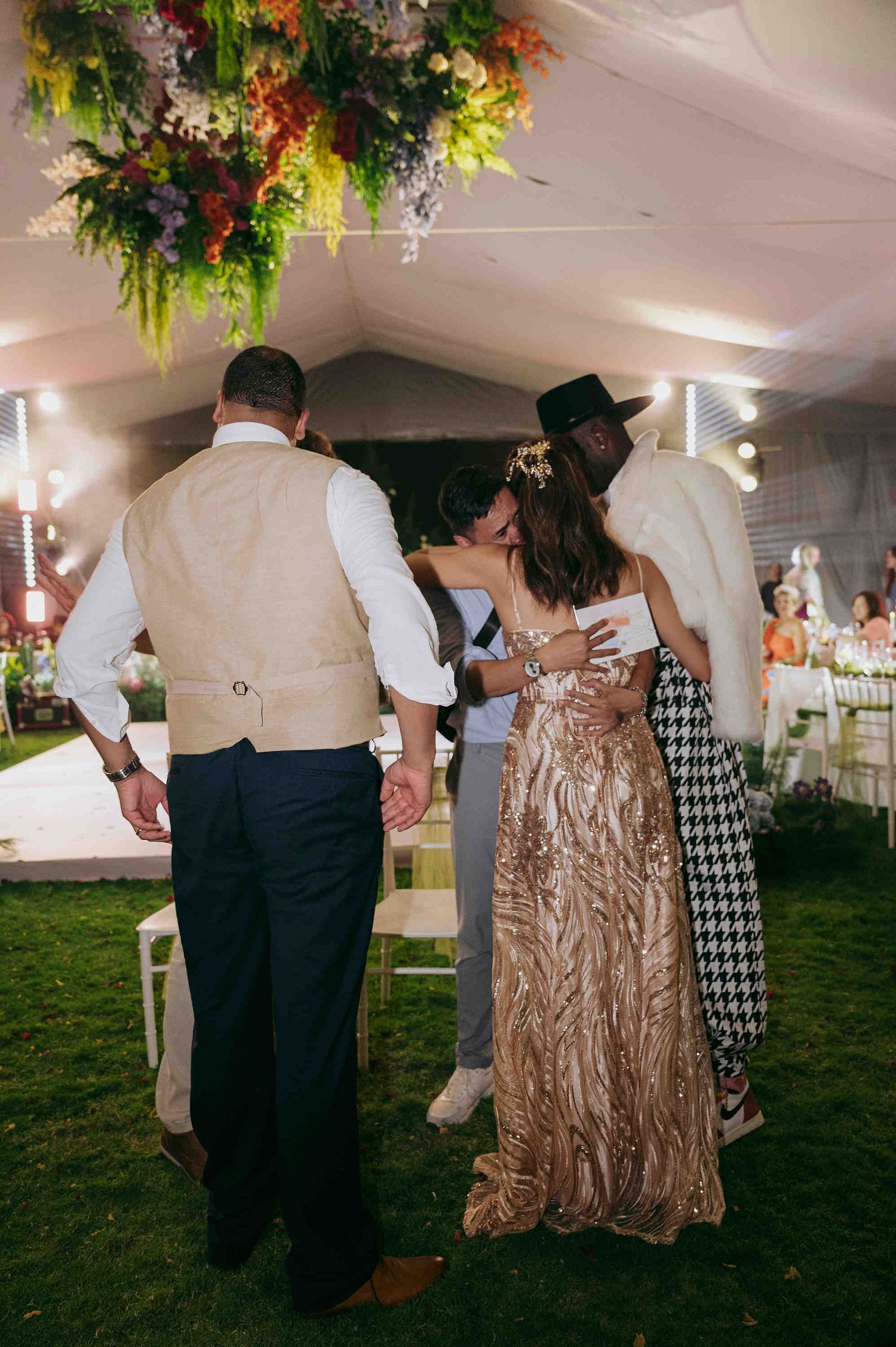 Guests in formal attire embrace at an outdoor wedding reception under floral arrangements and warm string lights.