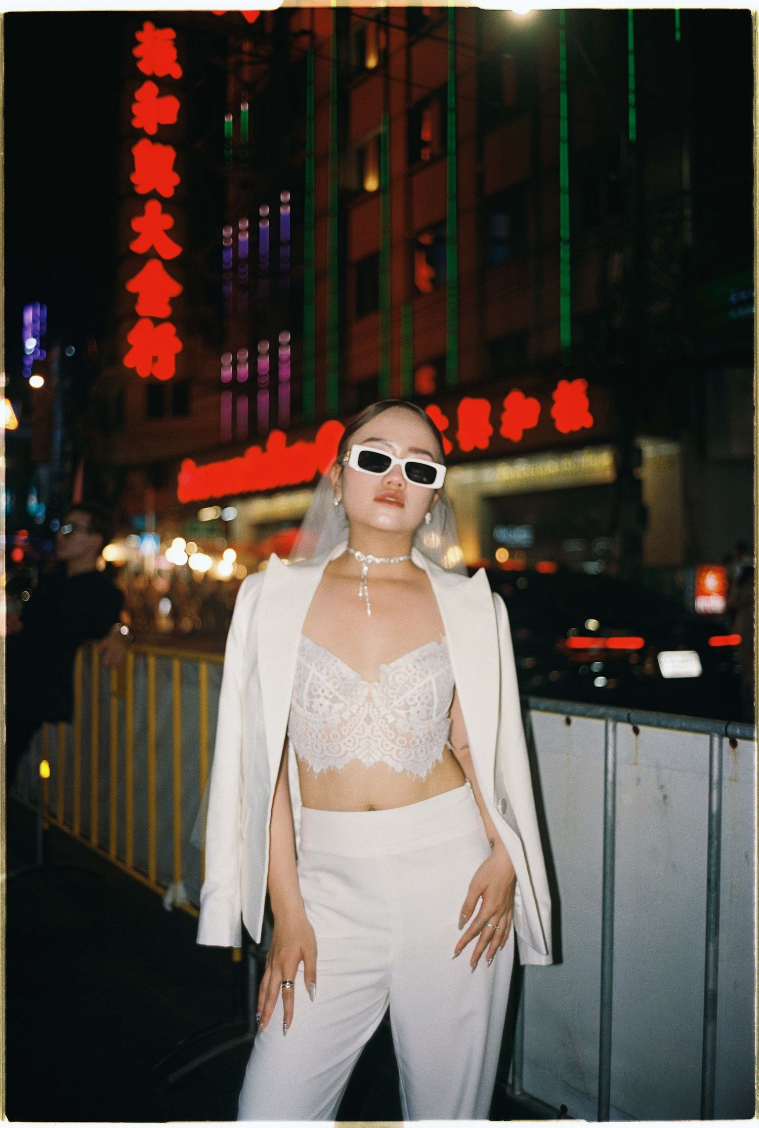 Saigon pre-wedding: person in white attire and sunglasses poses against metal barriers on a city street at night, by AnhVuWedding.