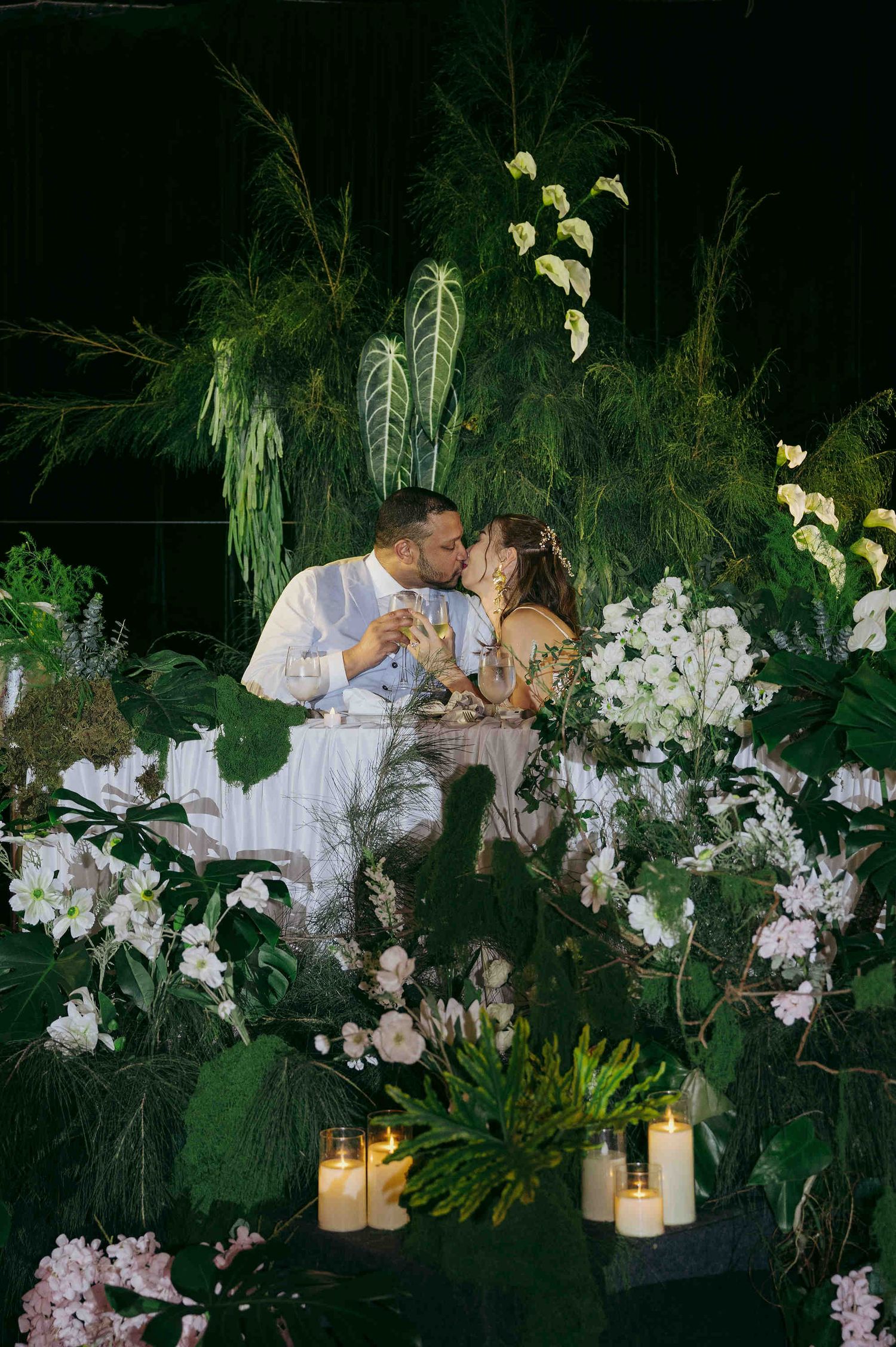 Couple sharing a romantic kiss surrounded by lush tropical greenery and white flowers with glowing candles at an elegant event.